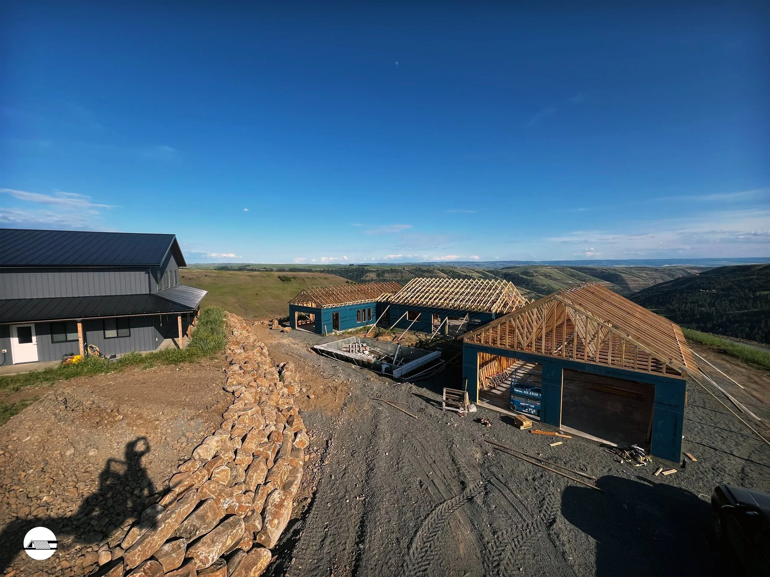Construction site of a building with a black metal roof, wooden framing for multiple rooms, and a rock-lined pathway, set in a rural landscape under a blue sky.