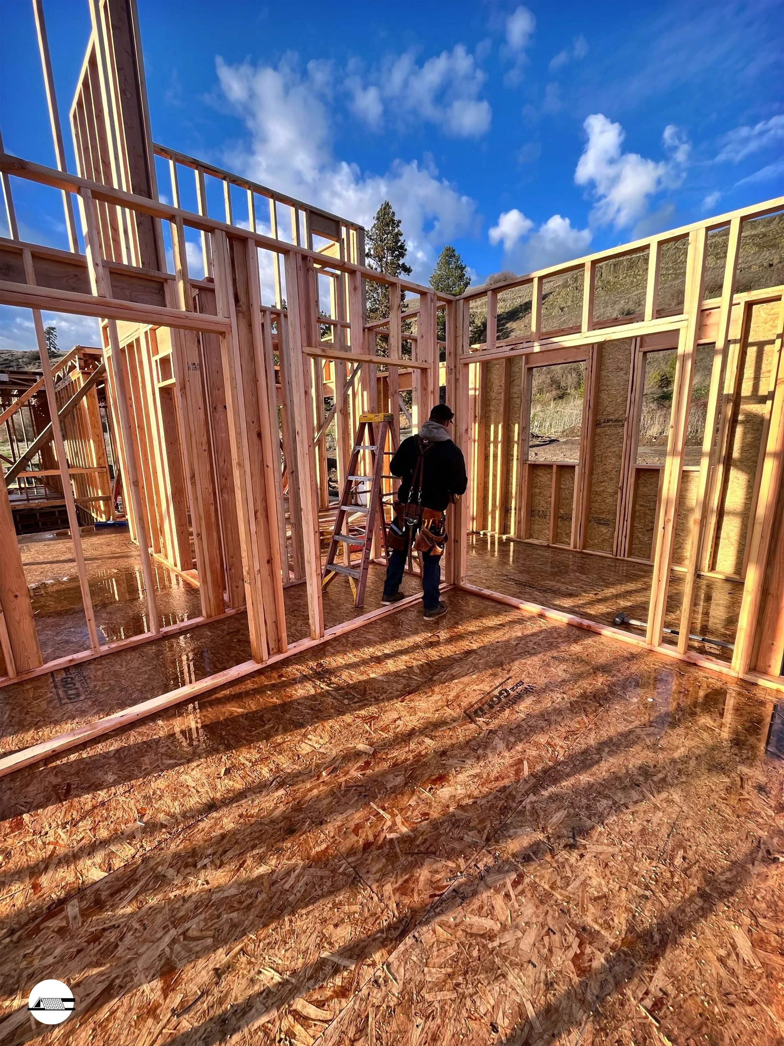 Construction site with framing of wooden walls and a worker standing near a ladder, under a blue sky with clouds.
