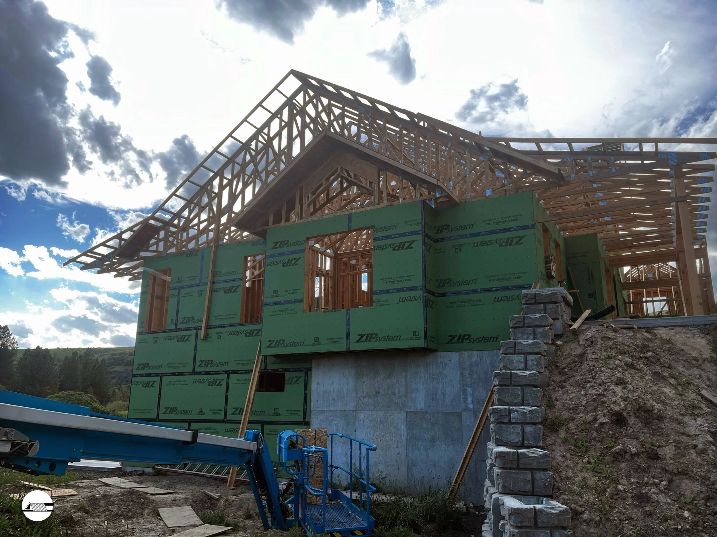 Construction site of a house with a wooden frame and green sheathing, situated on a concrete foundation, under a partly cloudy sky.