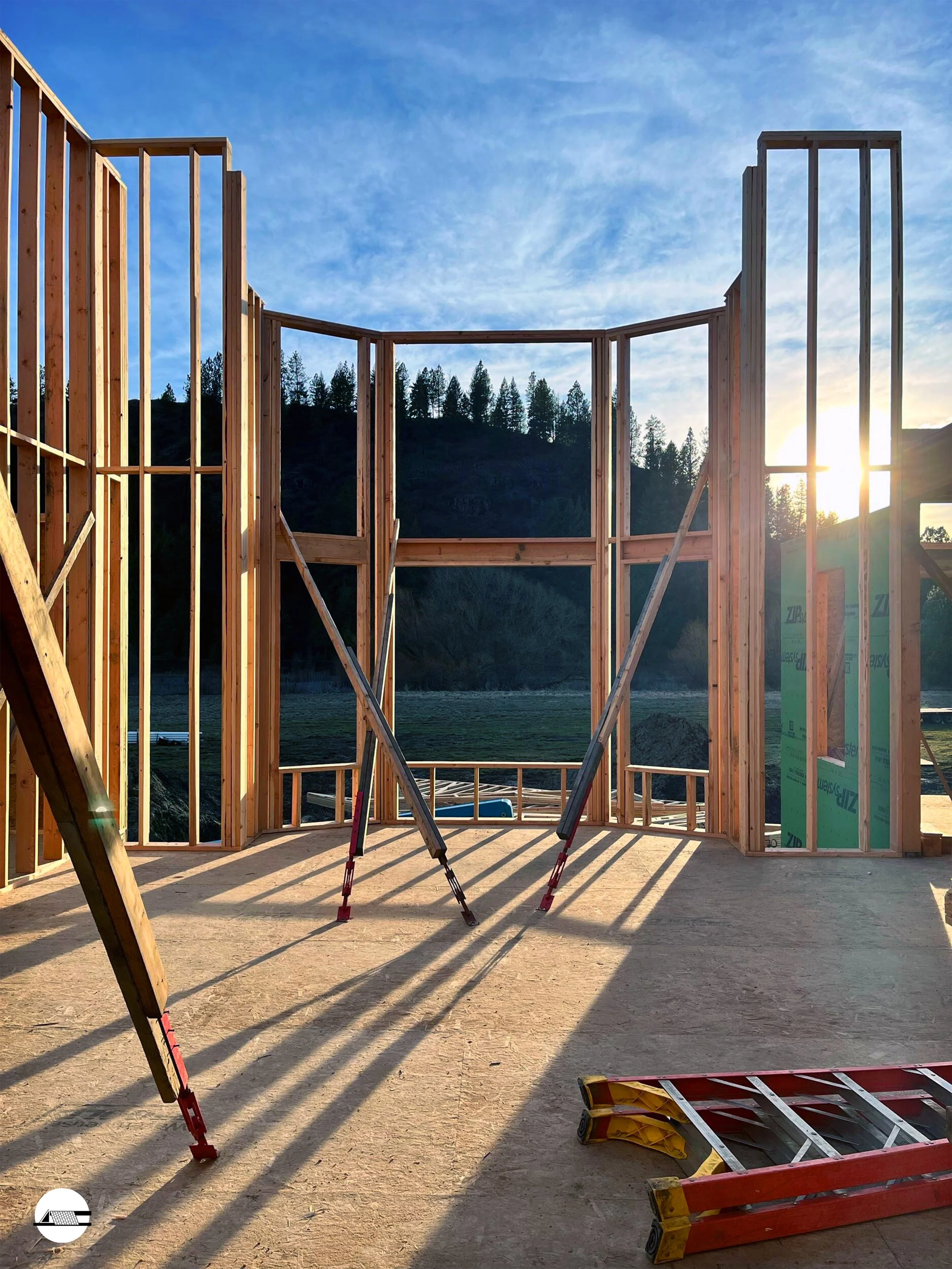 Wooden framing of a building under construction with open walls, sunlight shining through, and a ladder lying on the floor inside.