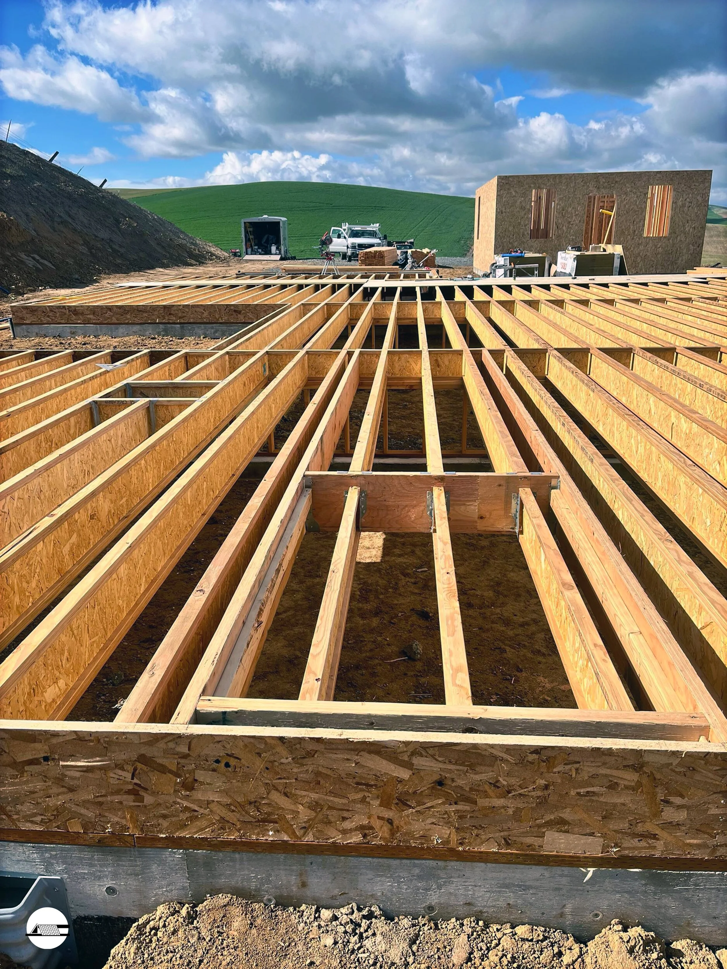 Construction site with wooden framing for a building floor, with a background of green rolling hills and cloudy sky.