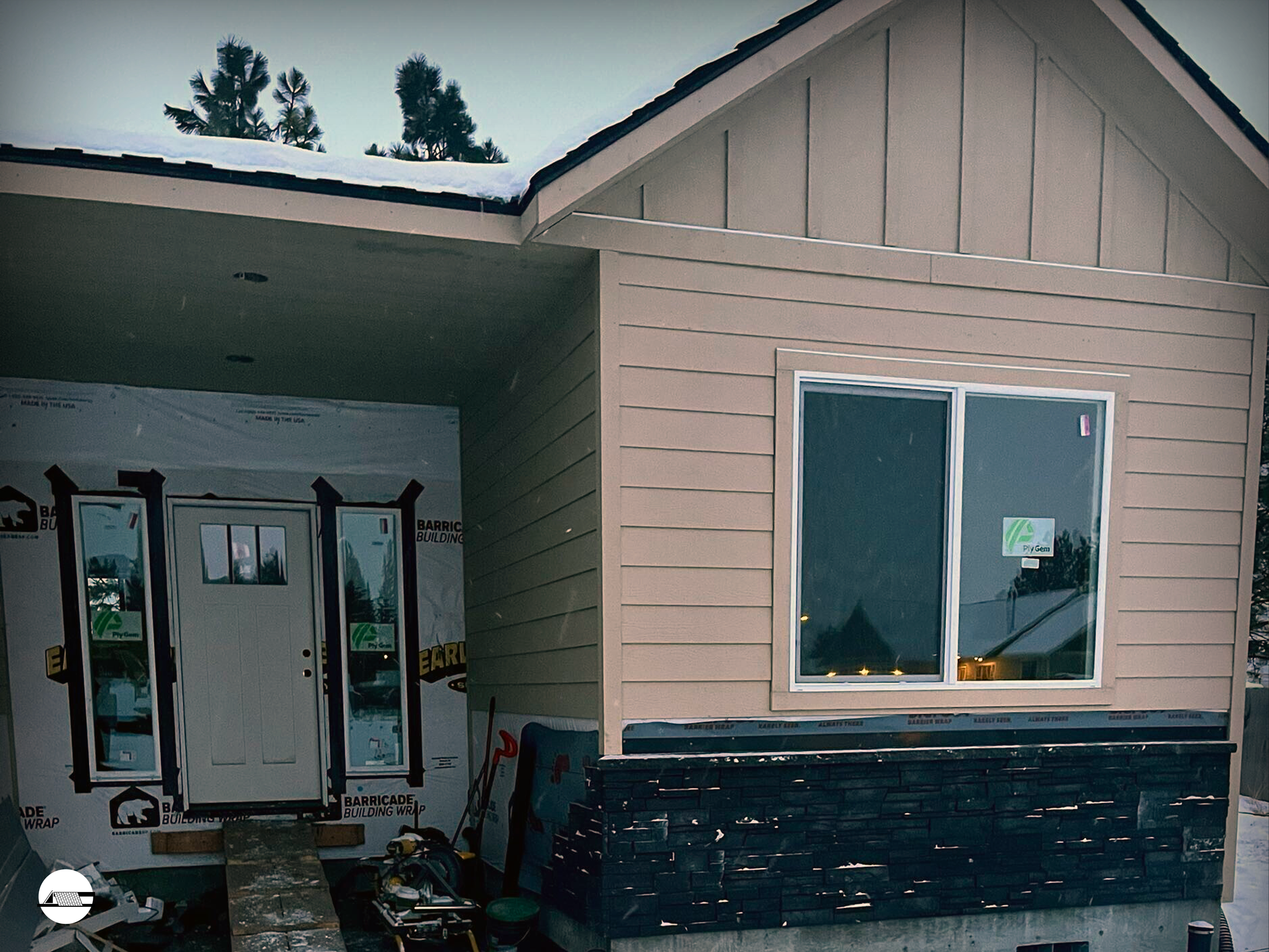 Front of a house under construction, with beige siding, a window, and a black stone accent at the base, with construction tools and materials nearby, and snow on the roof.