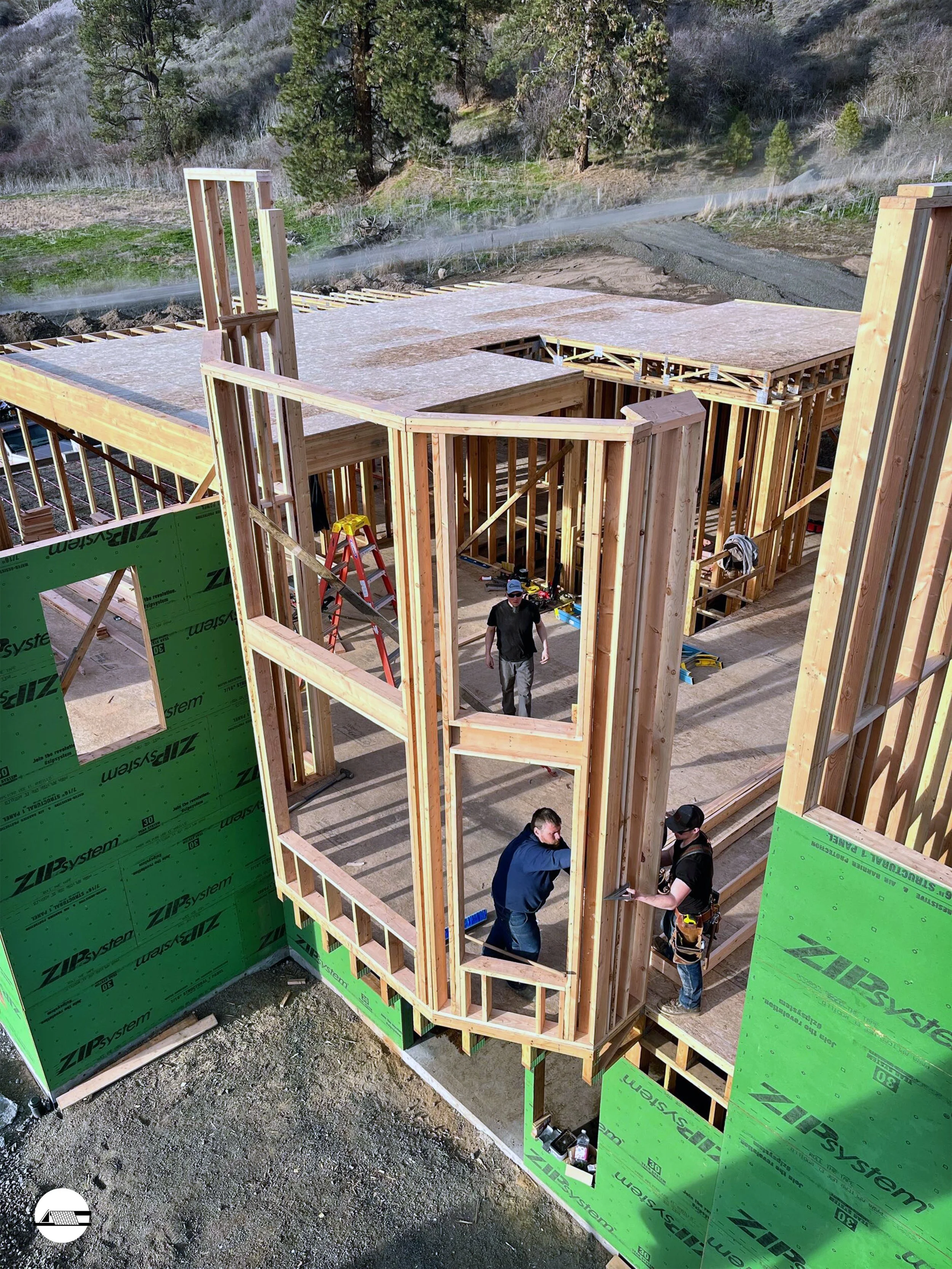 Construction workers building the wooden framework of a house in a rural area with trees and dirt roads in the background.