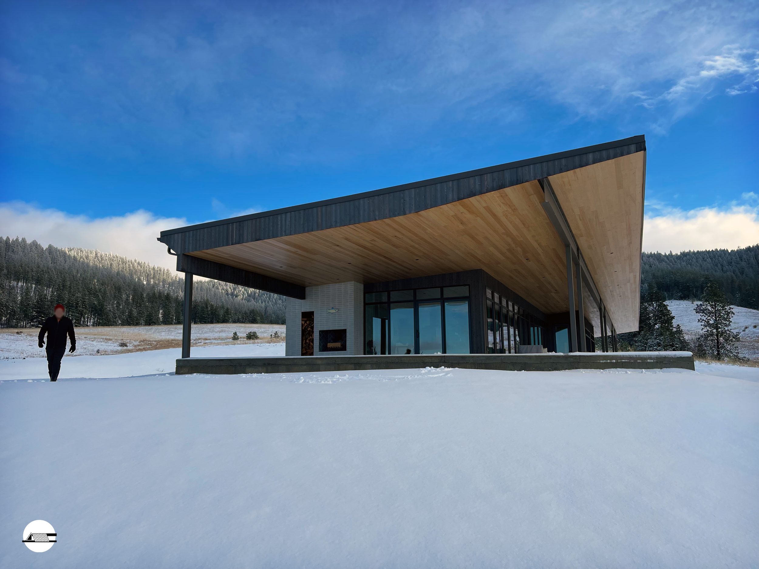 Modern house with a large wooden roof, glass walls, and a snow-covered landscape with a person walking nearby under a blue sky with clouds.