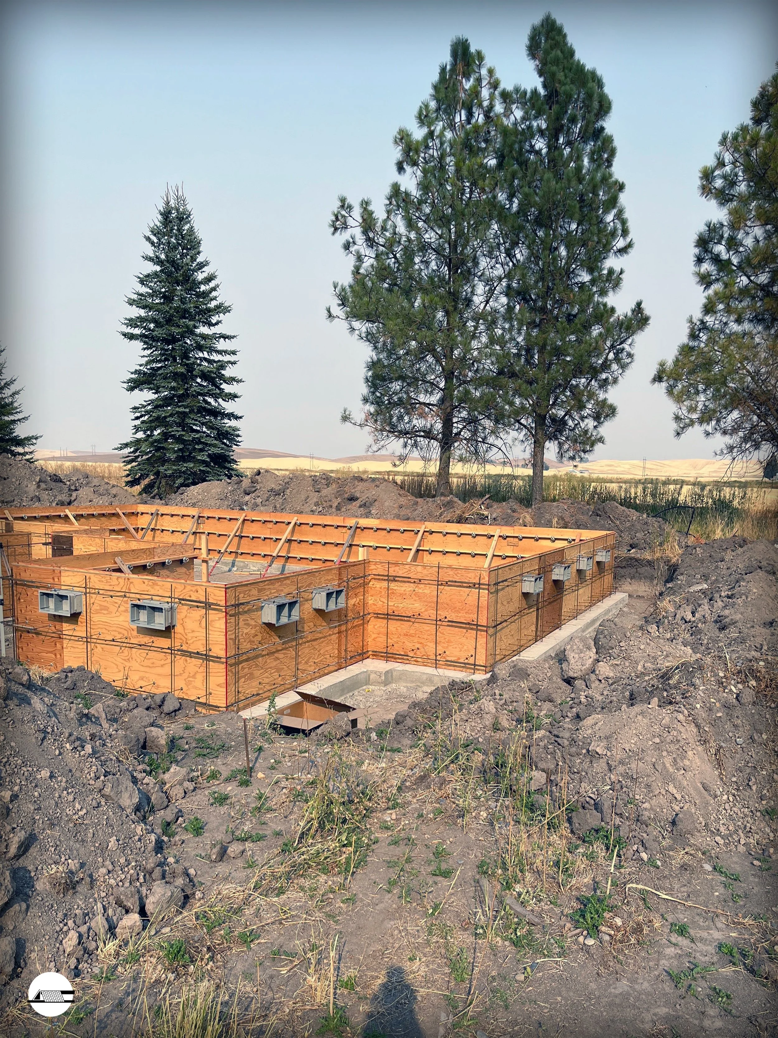 Construction site with wooden framing and concrete foundation, surrounded by dirt and trees in a rural area.