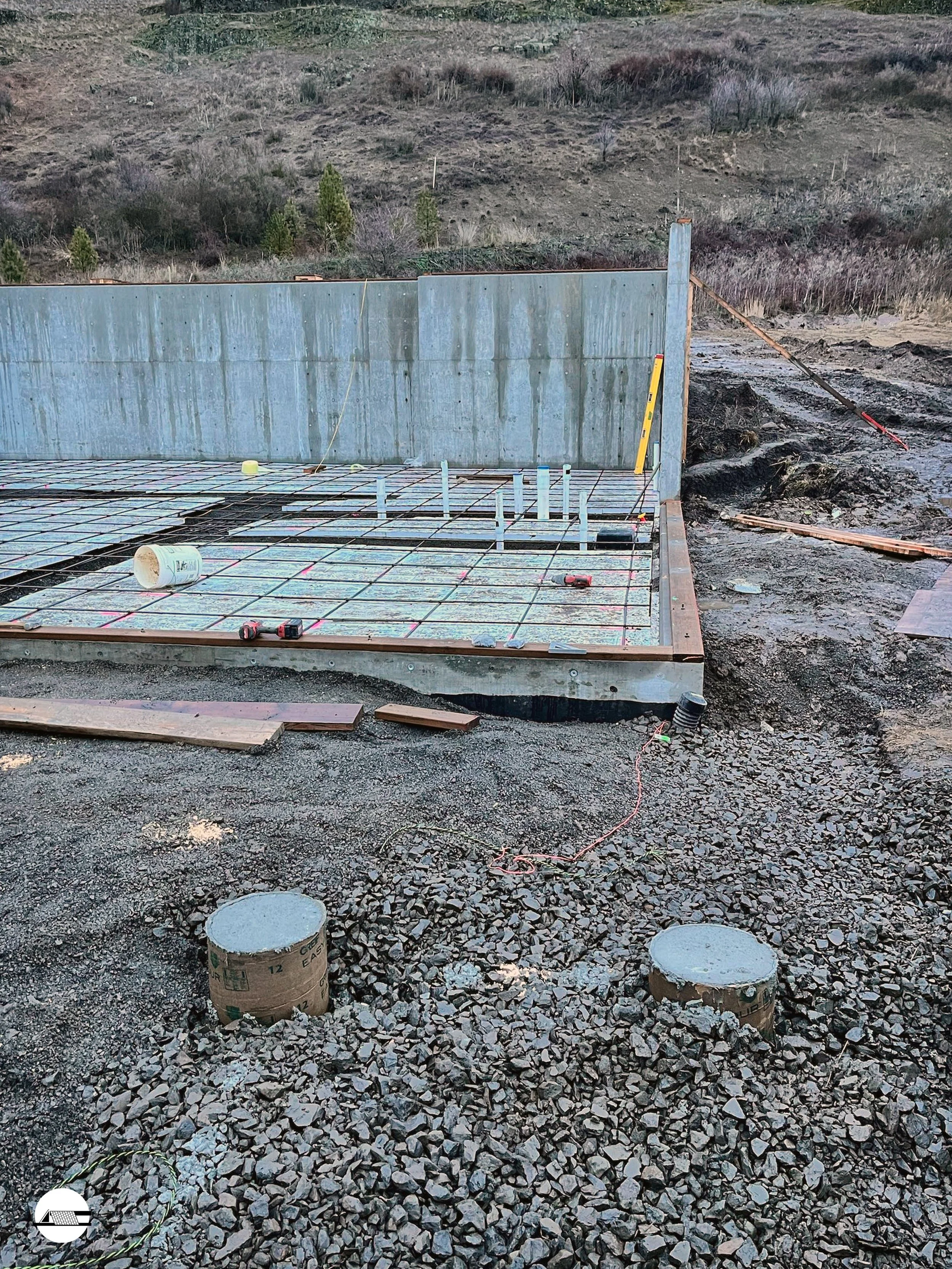 Construction site with concrete foundation and metal rebar grid, surrounded by dirt and gravel, with a hillside in the background.