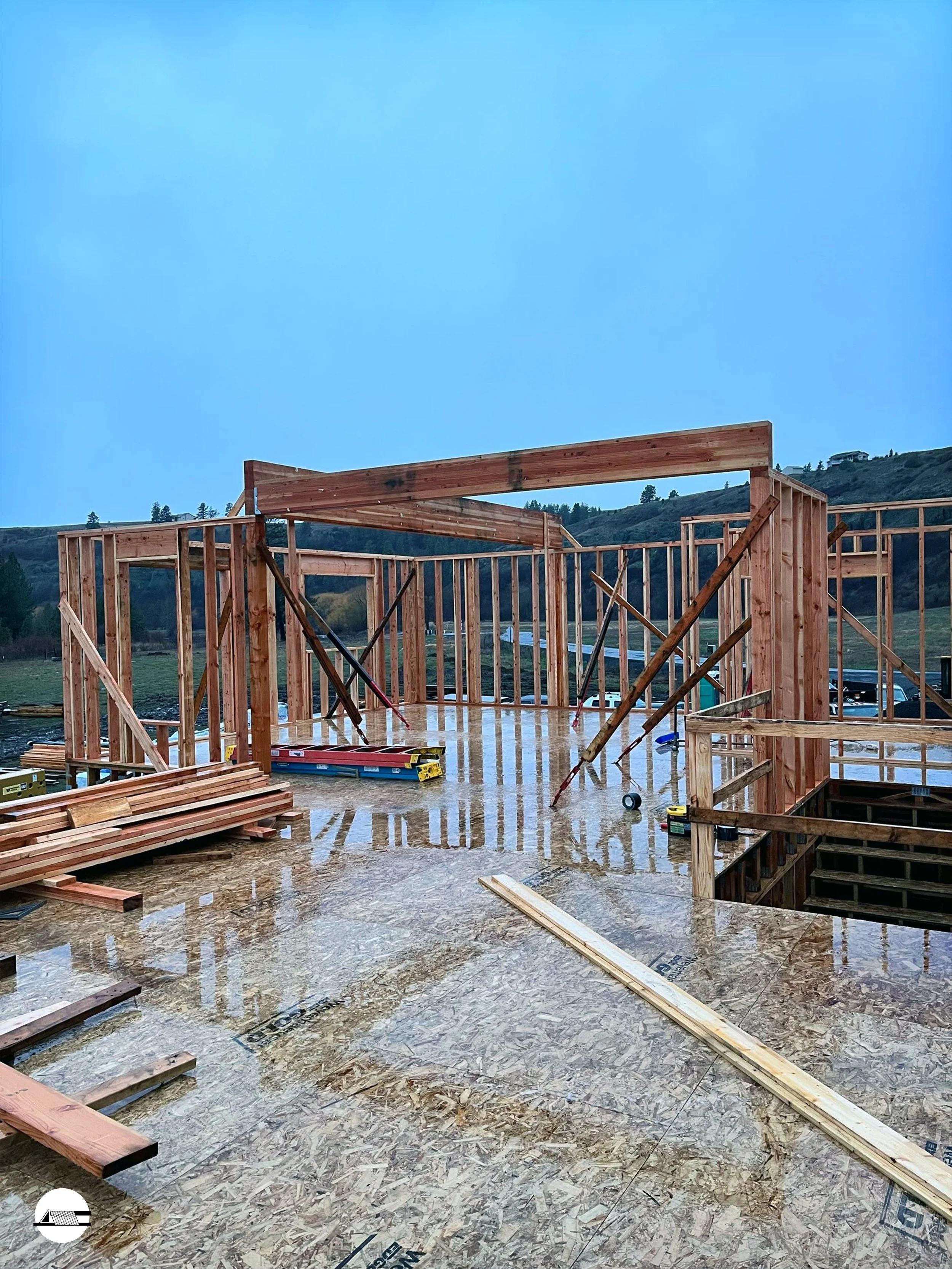 Construction site with wooden framing and plywood flooring, overcast sky, hills in background.