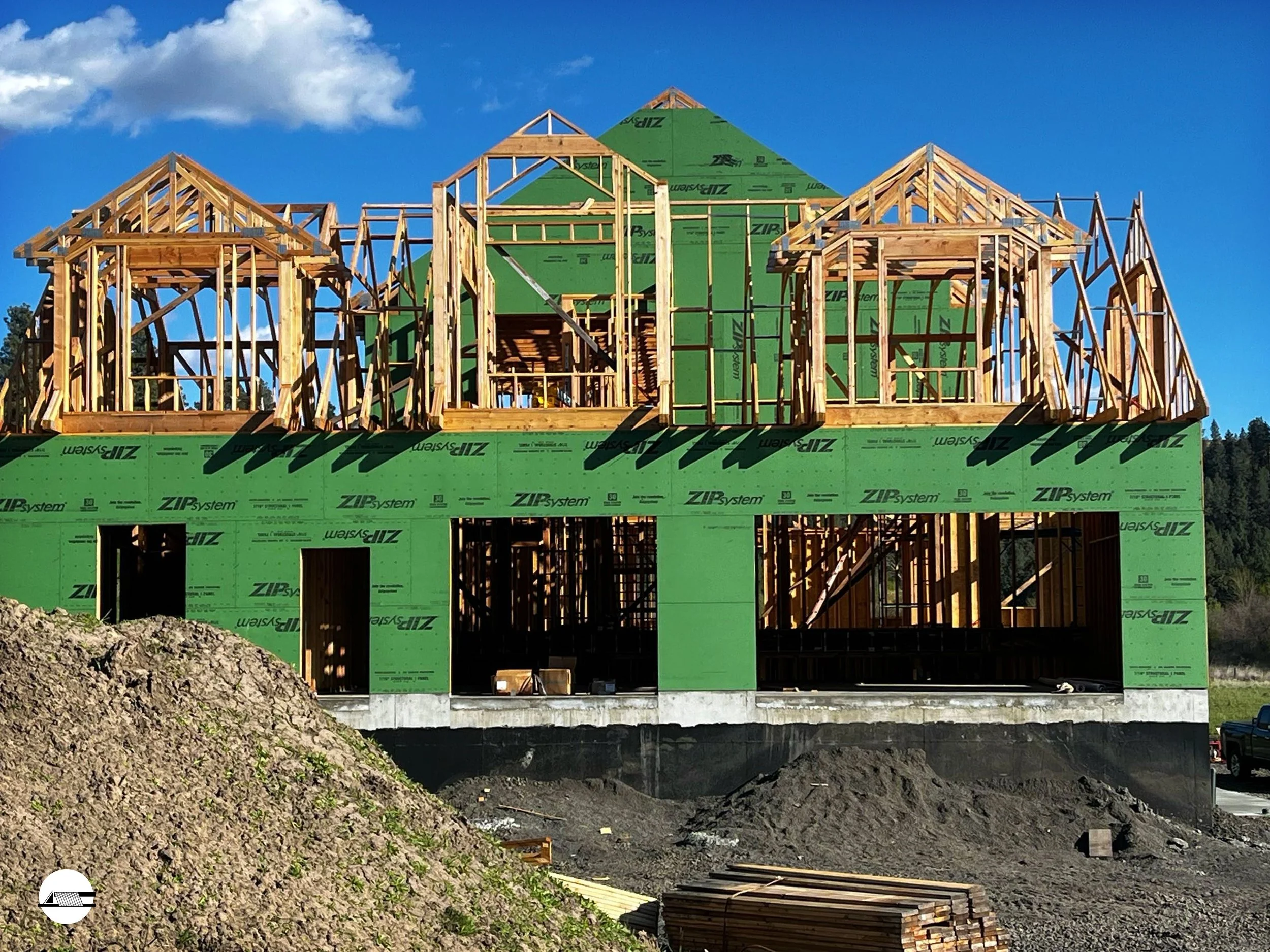 Construction site showing a partially built house with wooden framing and green sheathing, under a blue sky with some clouds.