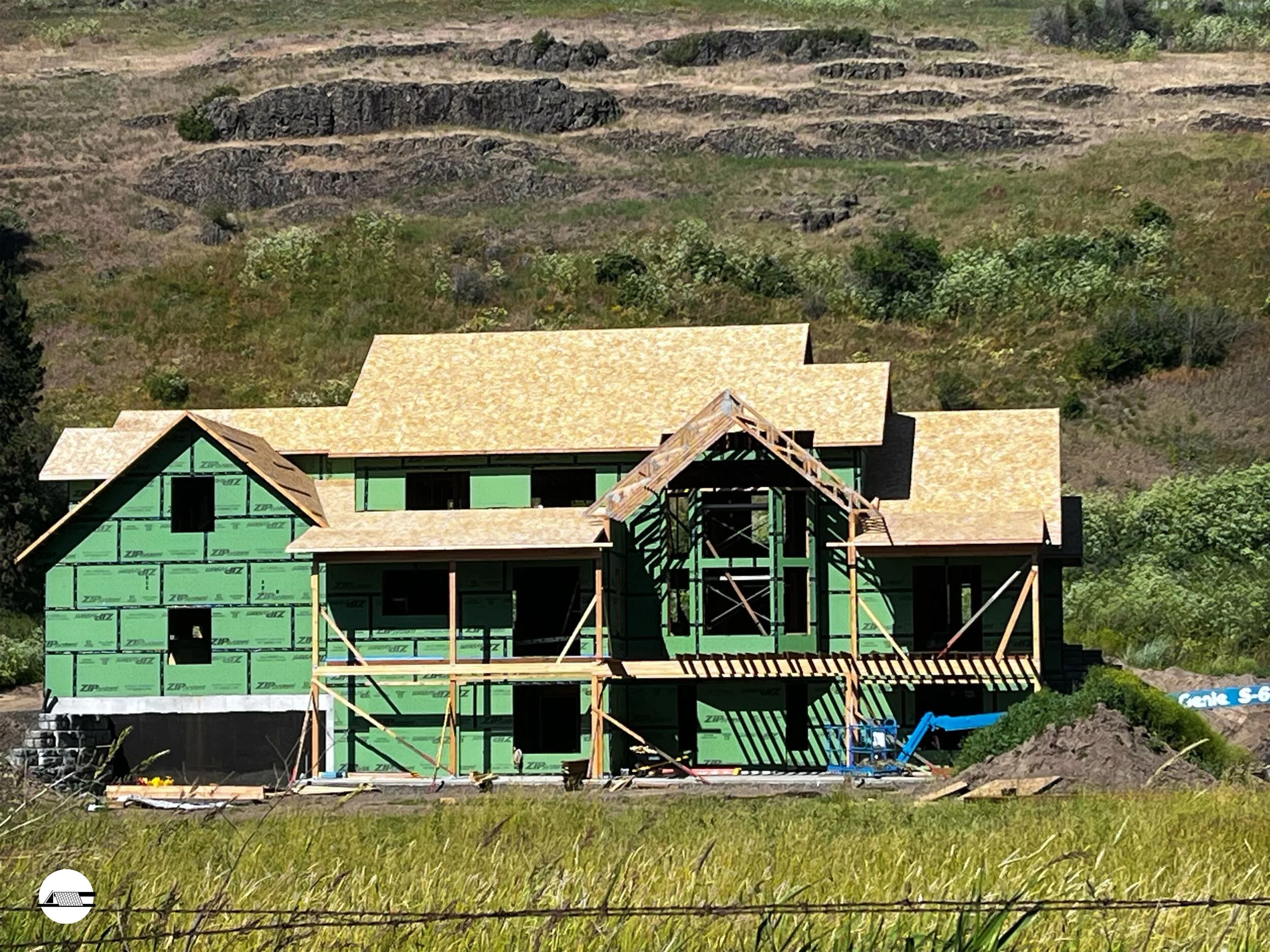 Under construction house with green sheathing, beige roof, wooden framing, and construction equipment in front, set against a hillside background.
