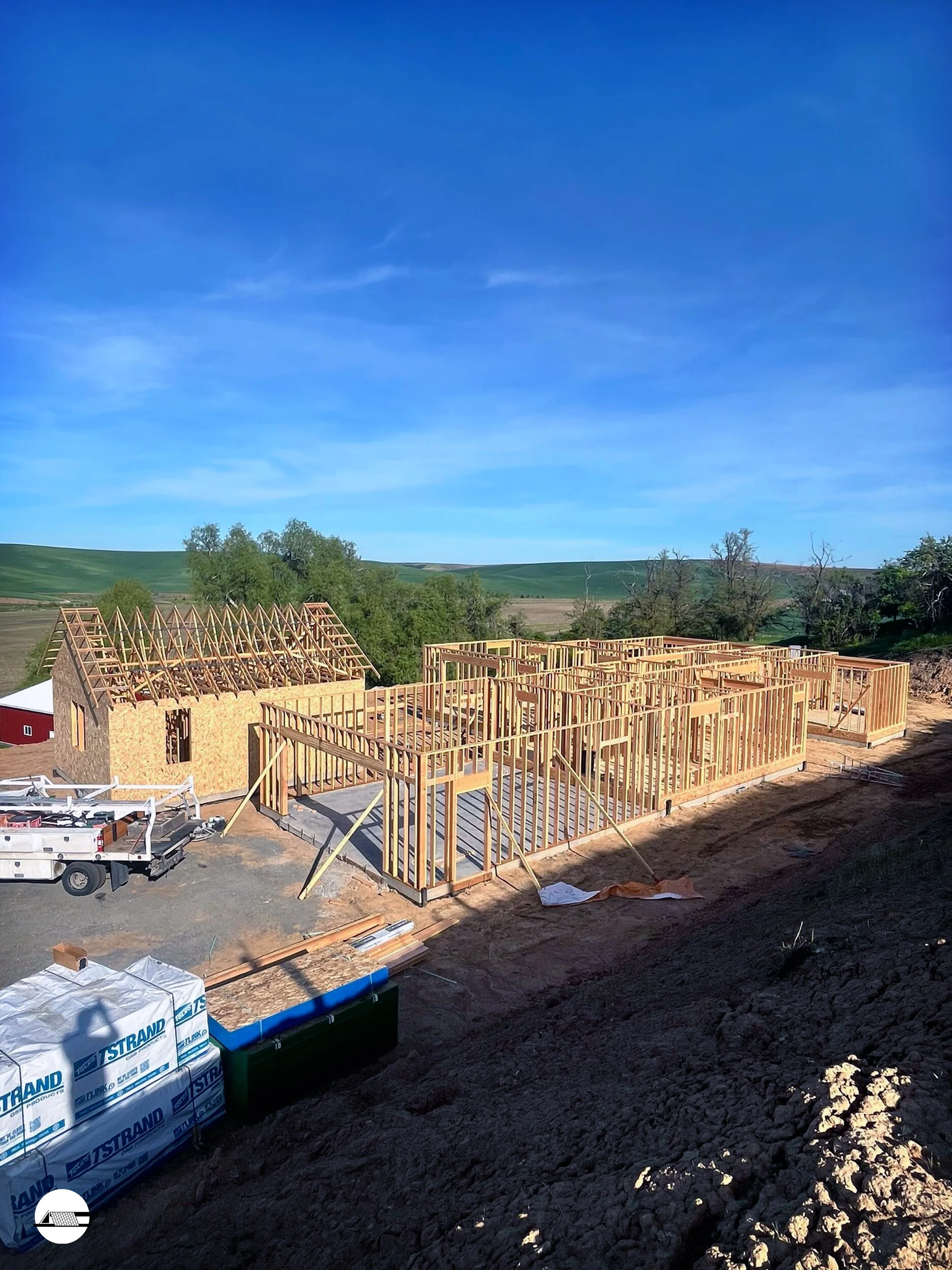 Wood framing for a house under construction with a clear blue sky and green landscape in the background.