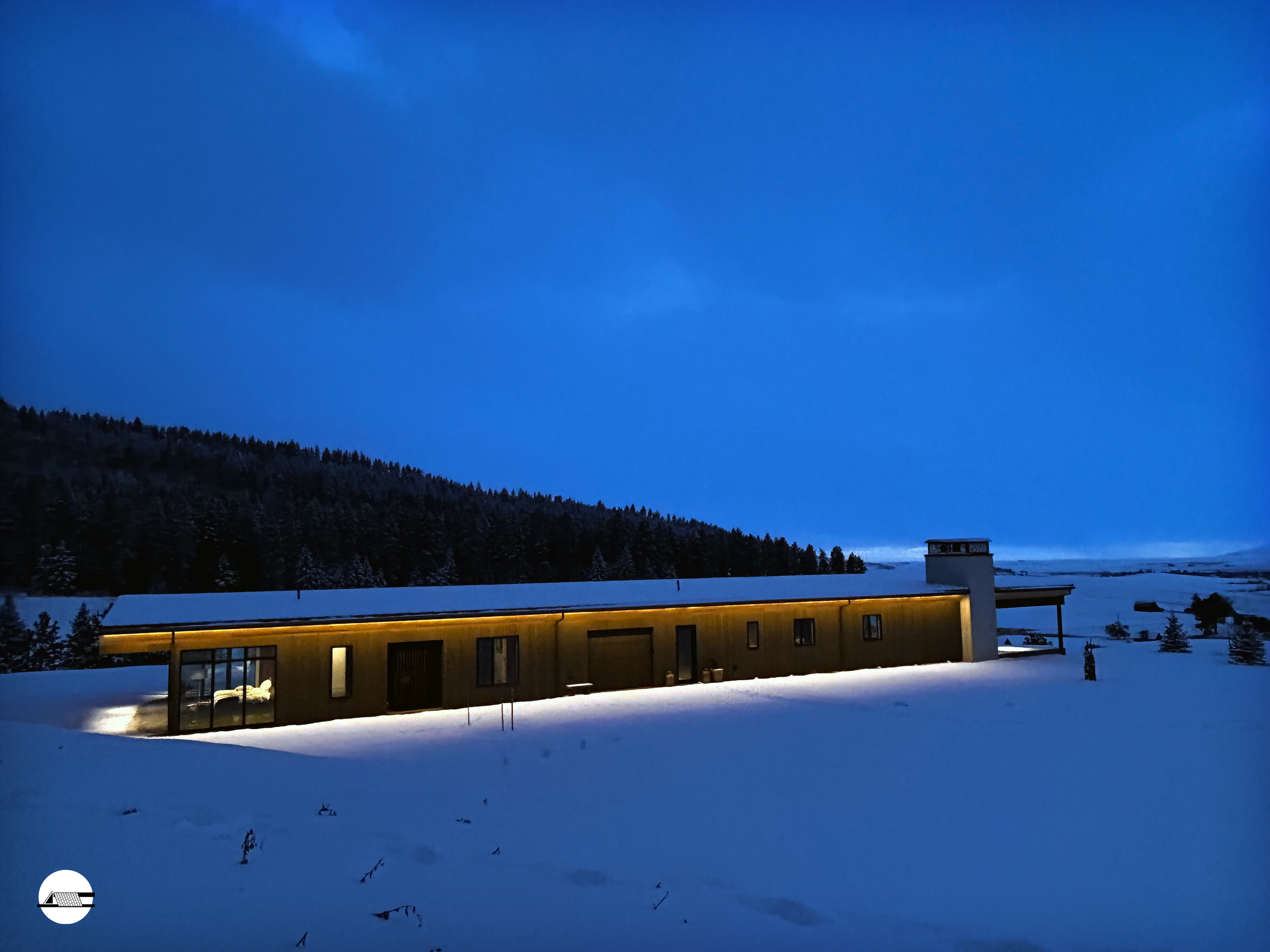Modern house with warm lighting in a snowy landscape under a dark blue twilight sky, surrounded by snow-covered trees and hills.