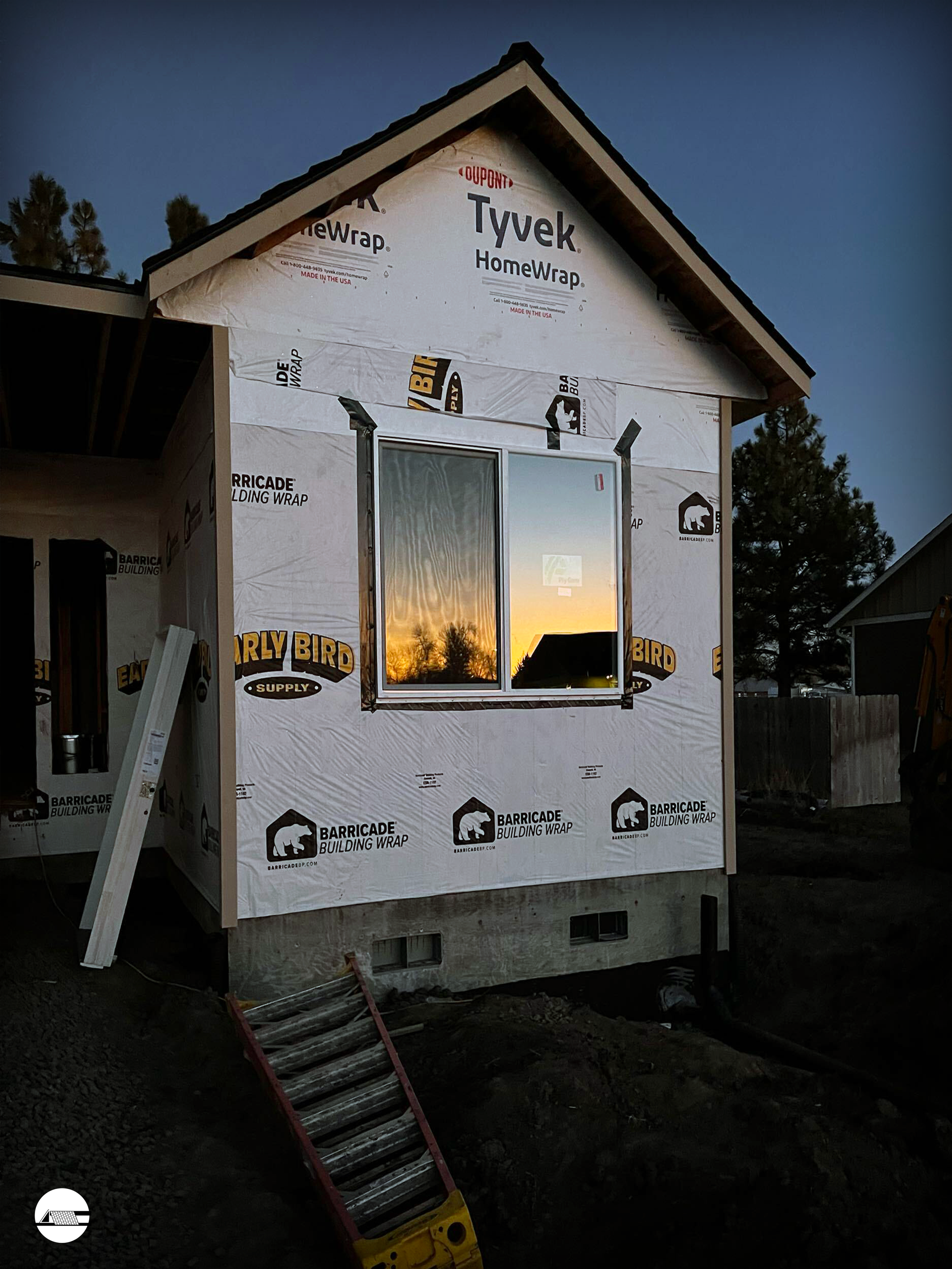 Corner of a house under construction with white weather-resistant siding wrap, a window reflecting a sunset, and a small ladder on the ground at dusk.