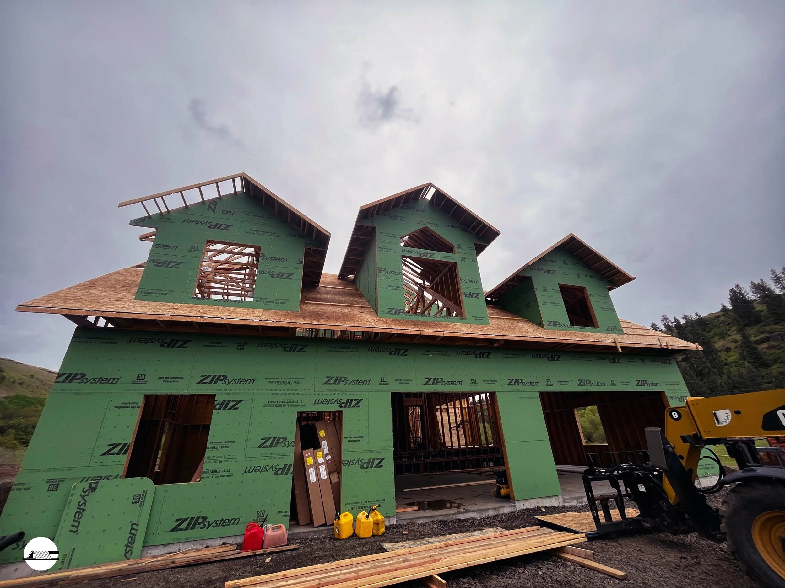 Under-construction house with green sheathing, multiple windows, and a garage, set against a hilly landscape with overcast sky.