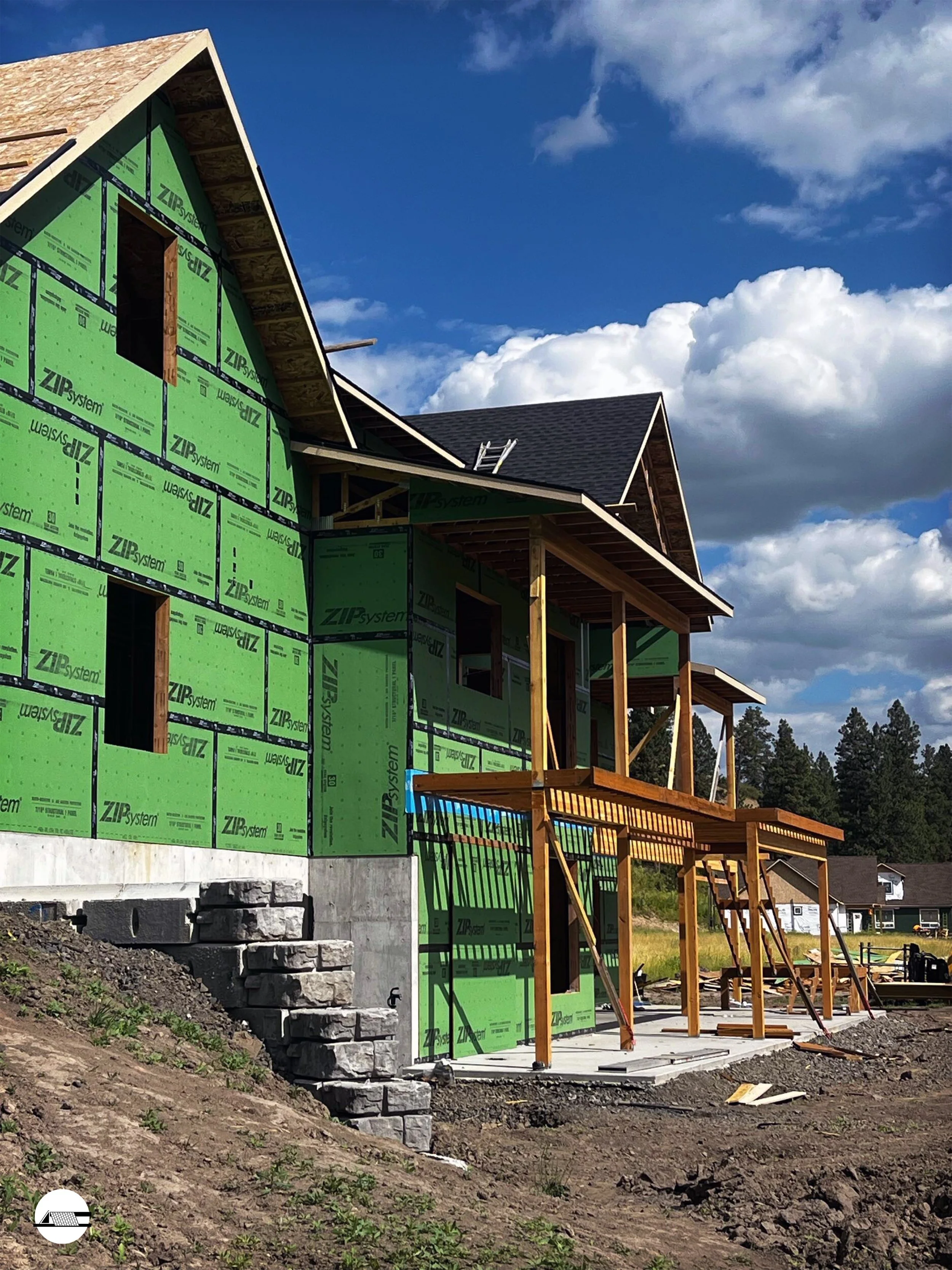 Under construction house with green sheathing and wooden framework in a residential area, clear sky with clouds in the background.