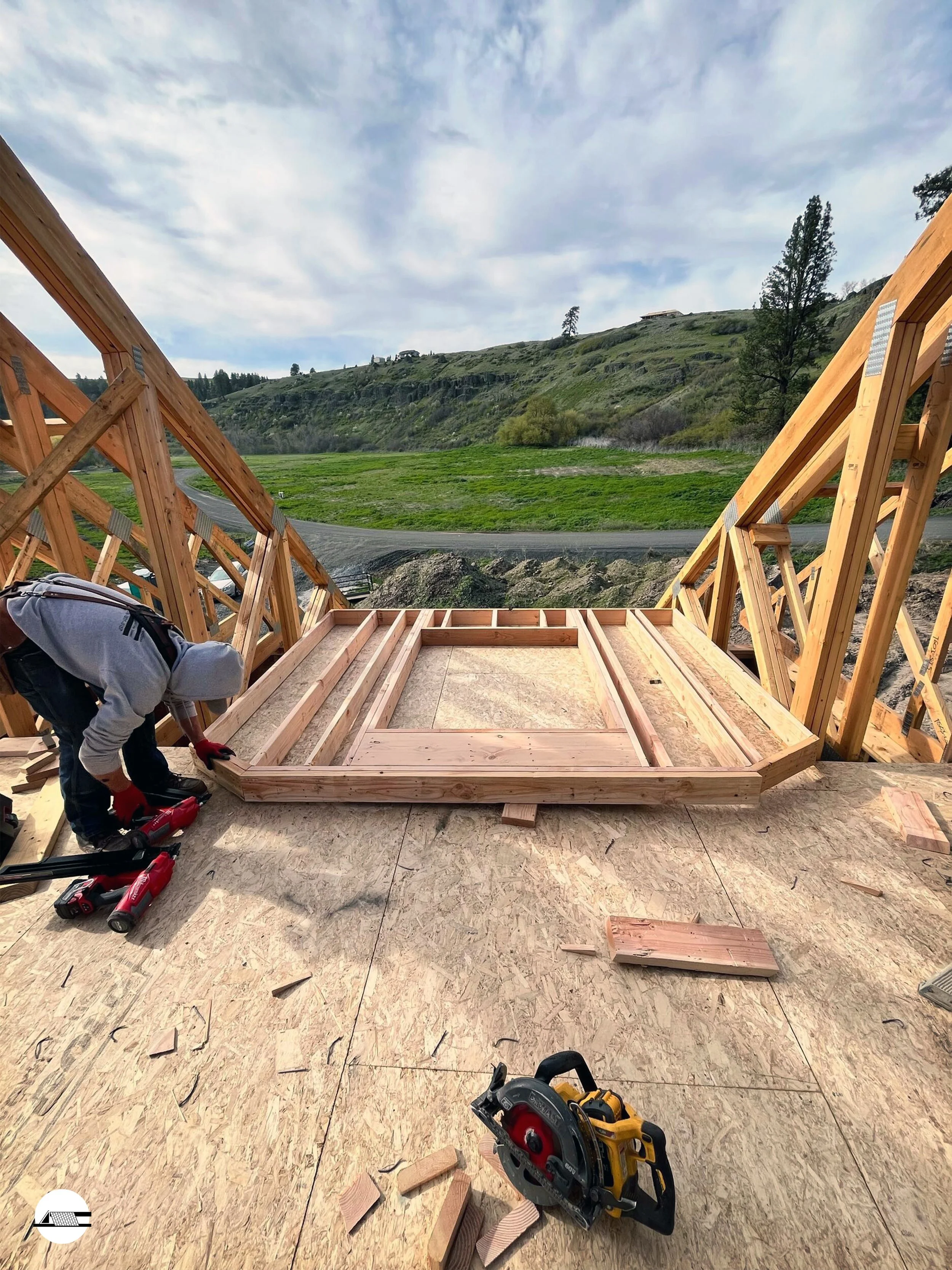 A construction worker building a wooden frame on a rooftop with scenic green hills and a cloudy sky in the background.