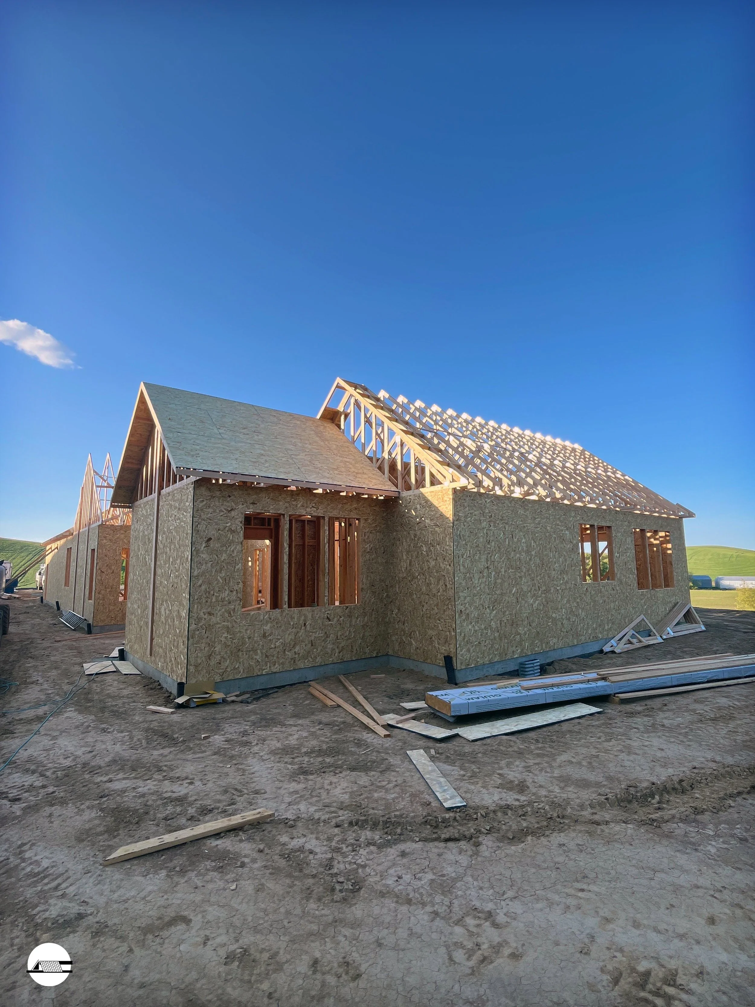 Construction site of a house with wooden framing and OSB sheathing, with the roof partially built, under a clear blue sky.