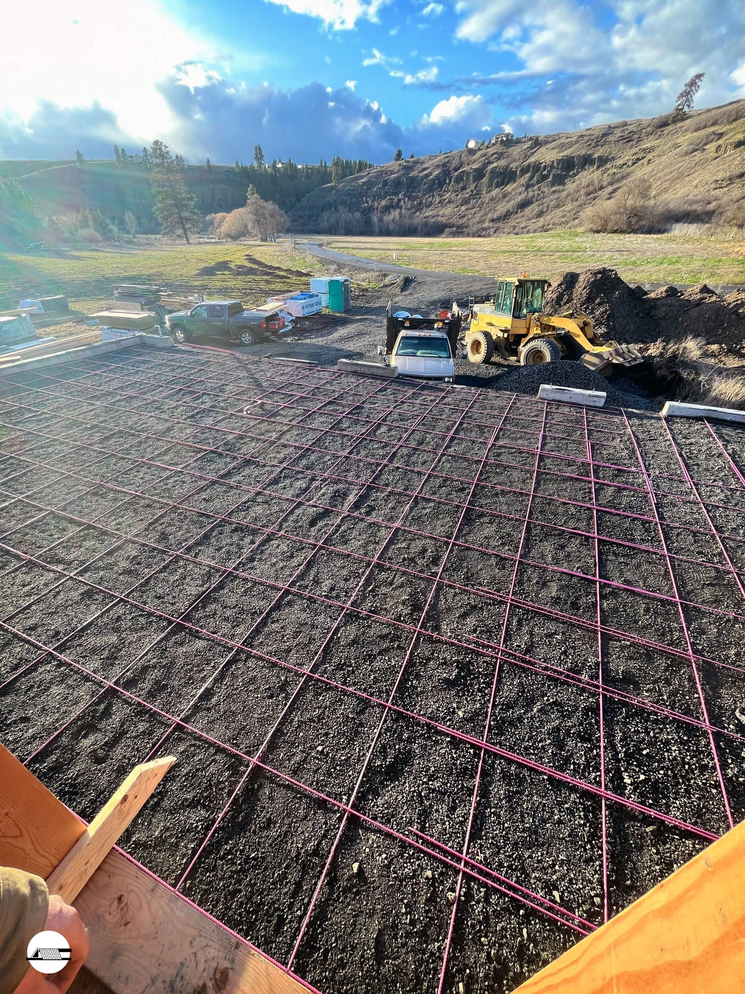 Construction site with a grid of pink rebar on dark soil, construction vehicles, and a hillside in the background under a partly cloudy sky.