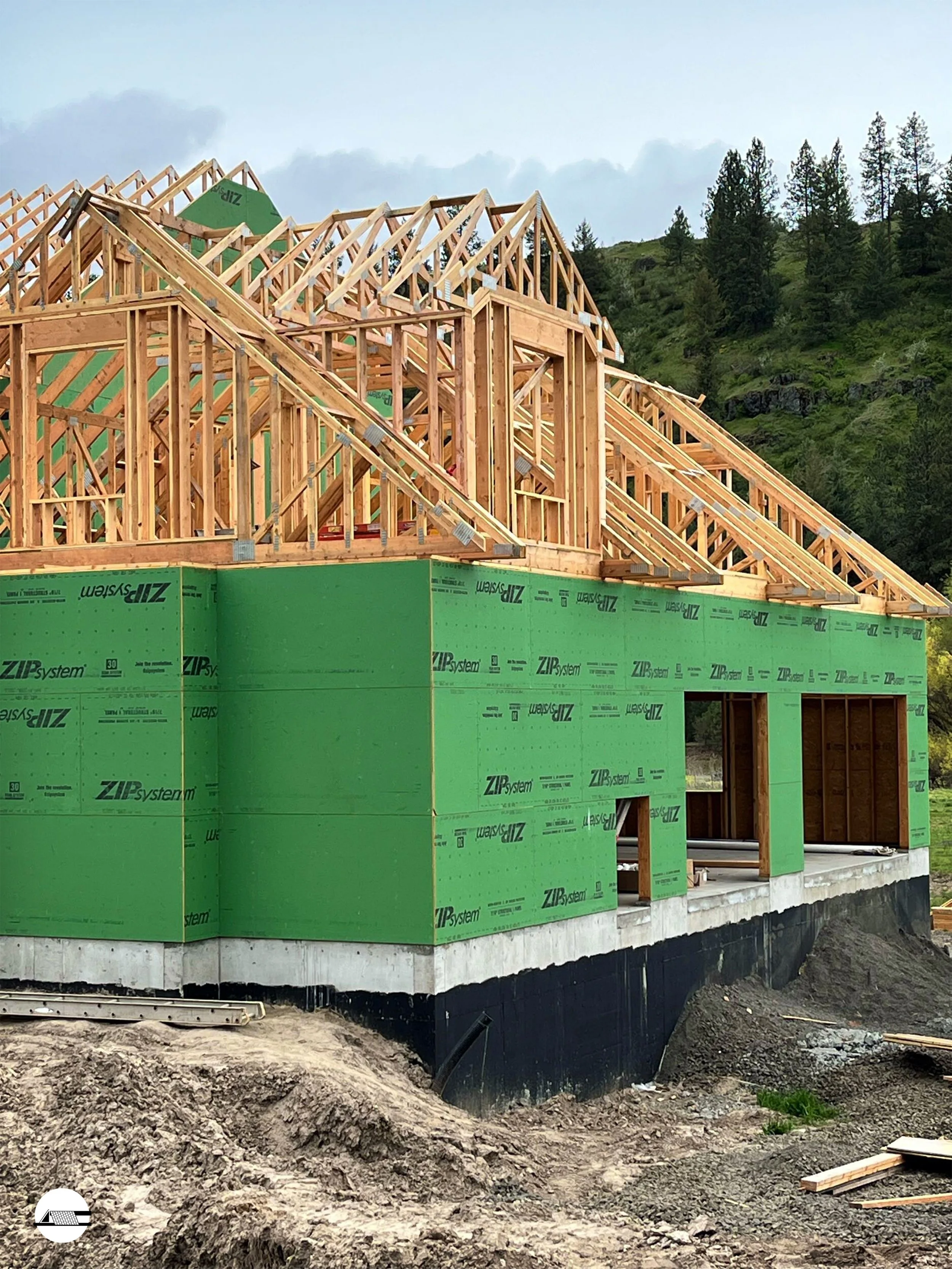 The image shows a house under construction with exposed wooden framing and green insulation panels on the exterior, situated on a sloped area with a forested hillside in the background.