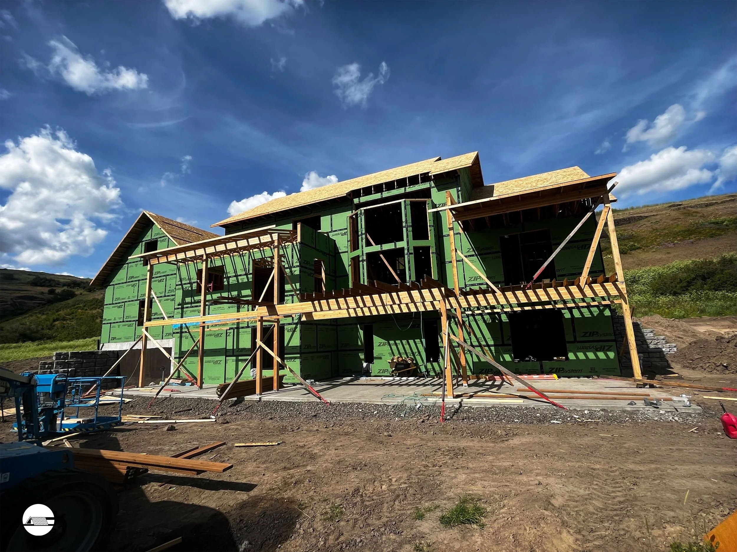 Under construction house with green sheathing, wooden framing, and scaffolding on a dirt lot, against a backdrop of mountains and blue sky with scattered clouds.