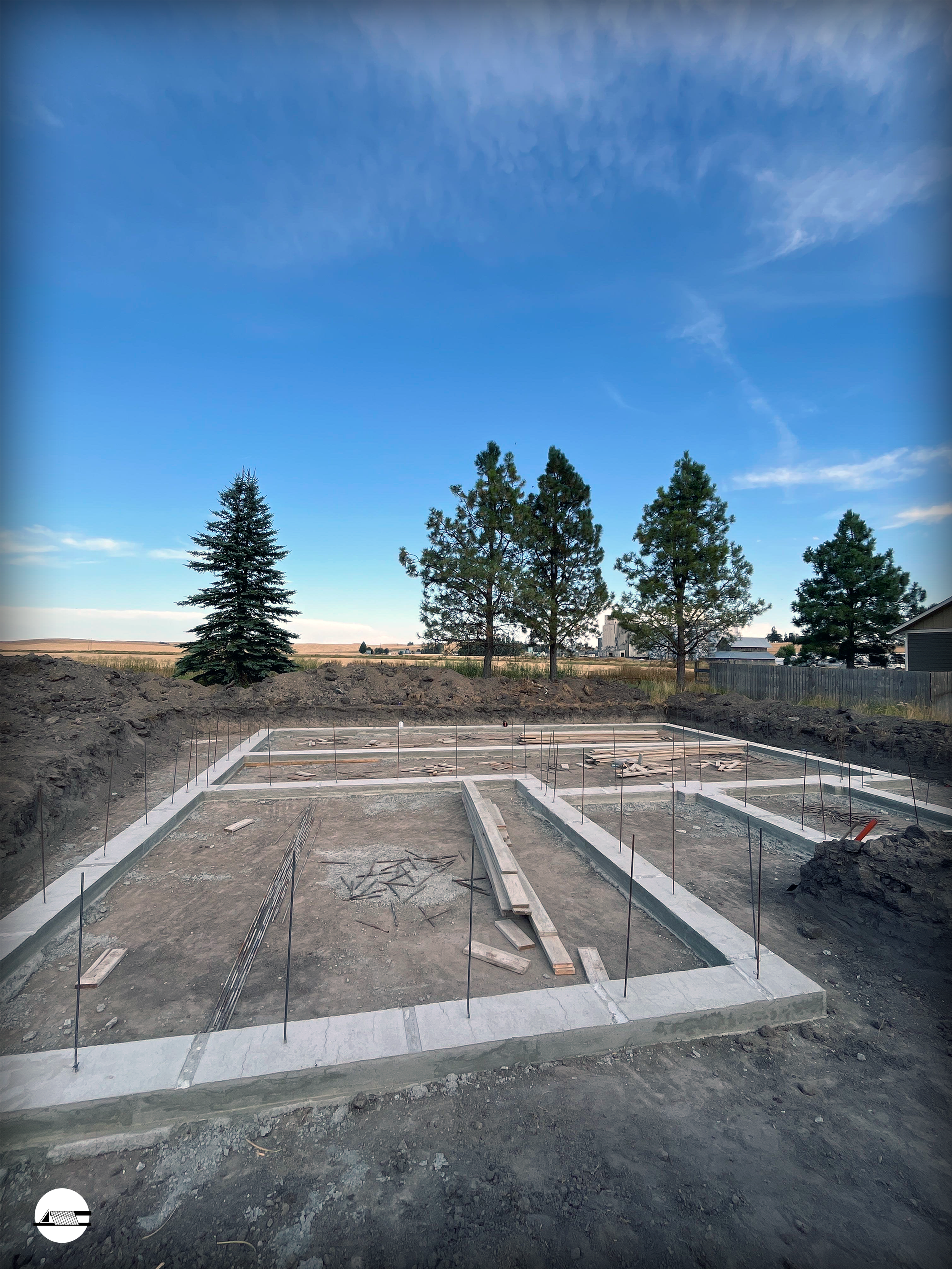 Concrete foundation for a building under construction, with wooden stakes and rebar, in a rural area with trees and an open sky.