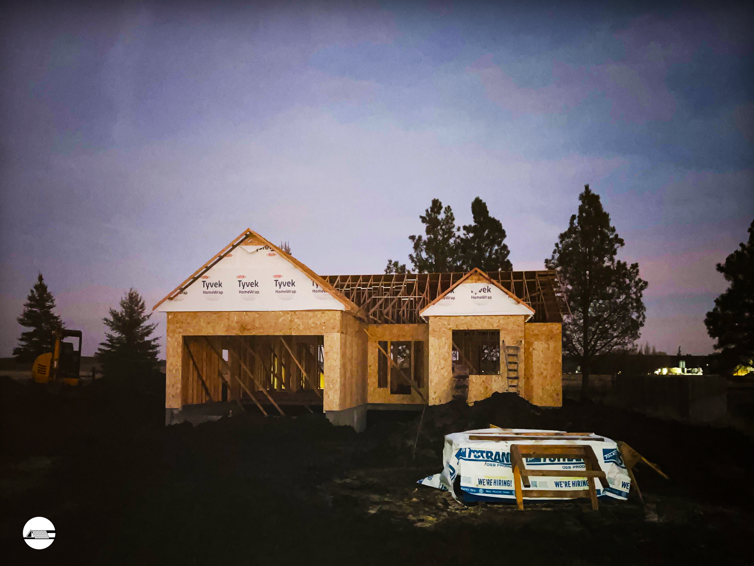 A house under construction with wooden framing and insulation in progress, set against a twilight sky with trees in the background.