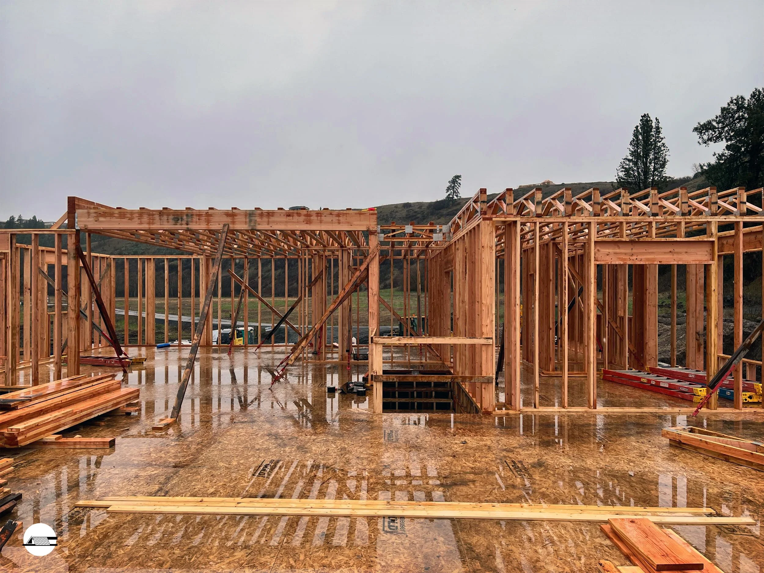 Wood framing of a house under construction on a rainy day, with visible wooden beams and plywood subfloor.