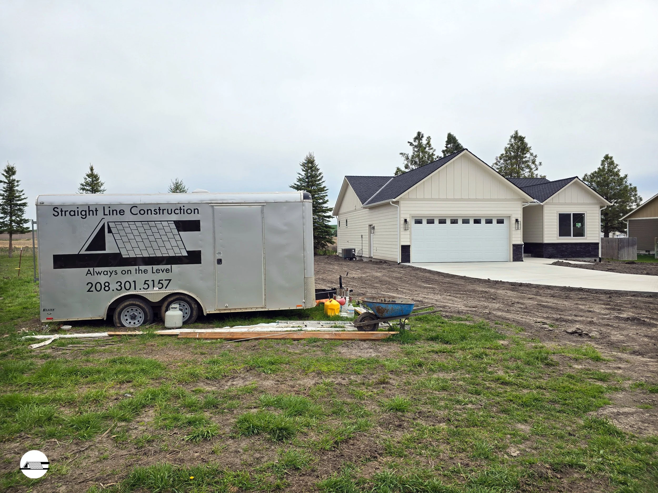 Newly constructed house with a white garage door, beige siding, a black shingled roof, and a concrete driveway. Construction trailer with the logo 'Straight Line Construction' and phone number, along with construction tools and materials in the foreg