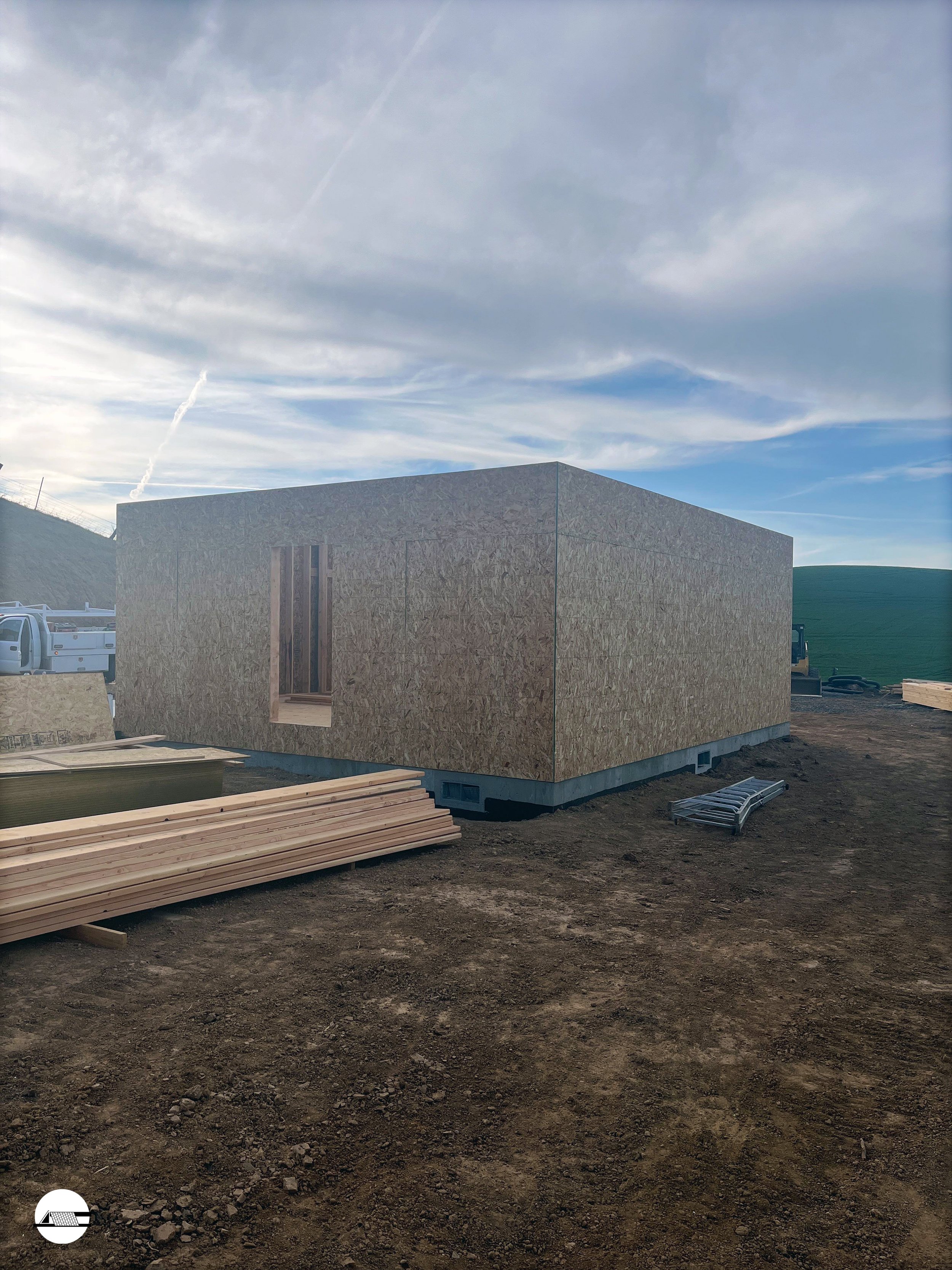Construction site featuring a small building under construction, with plywood walls, a slotted window opening, and construction materials like wood planks and metal grates on the ground.