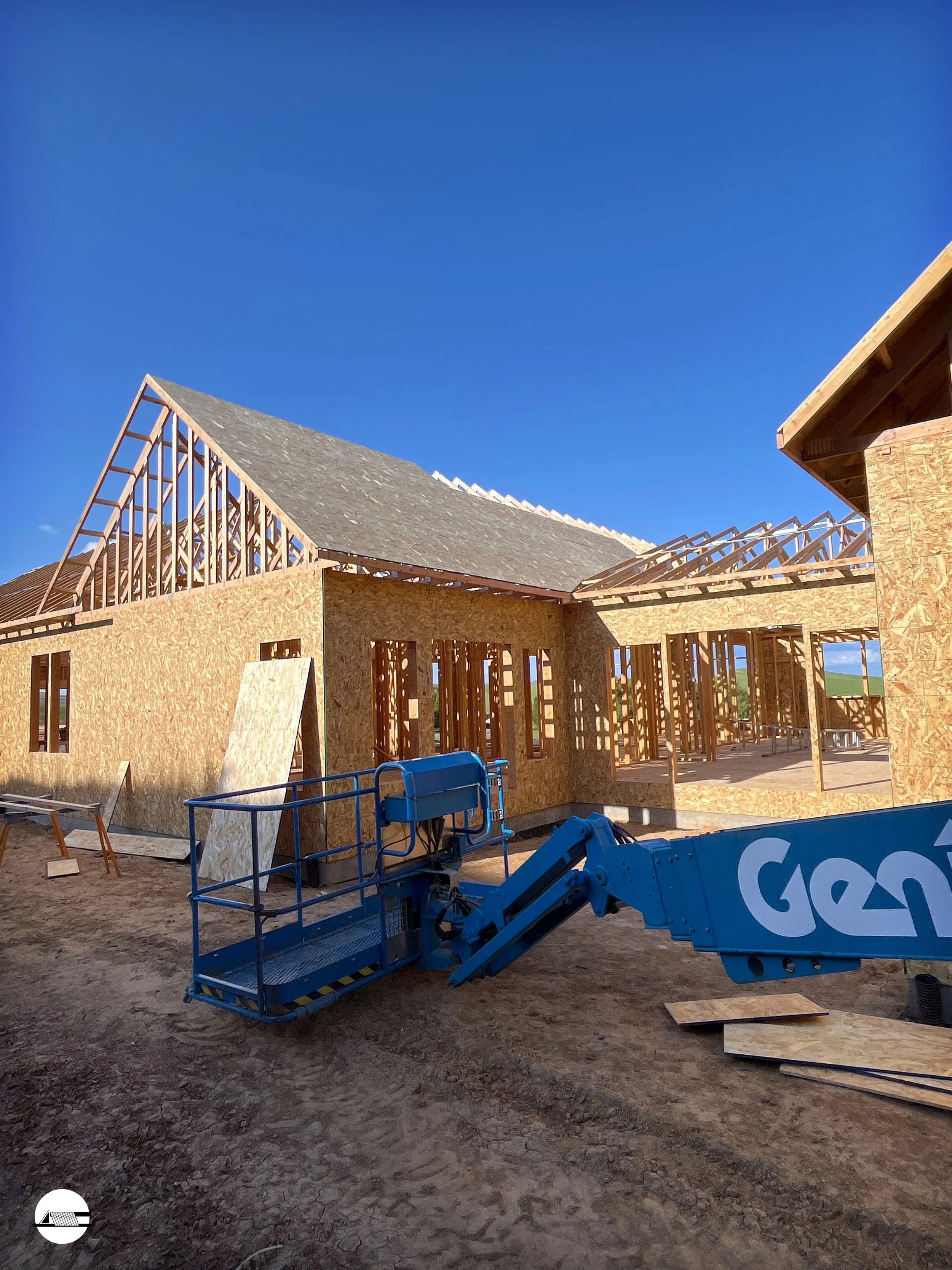 Construction site with wooden framing for a house, partially built, under a clear blue sky, with a blue lift equipment in the foreground.