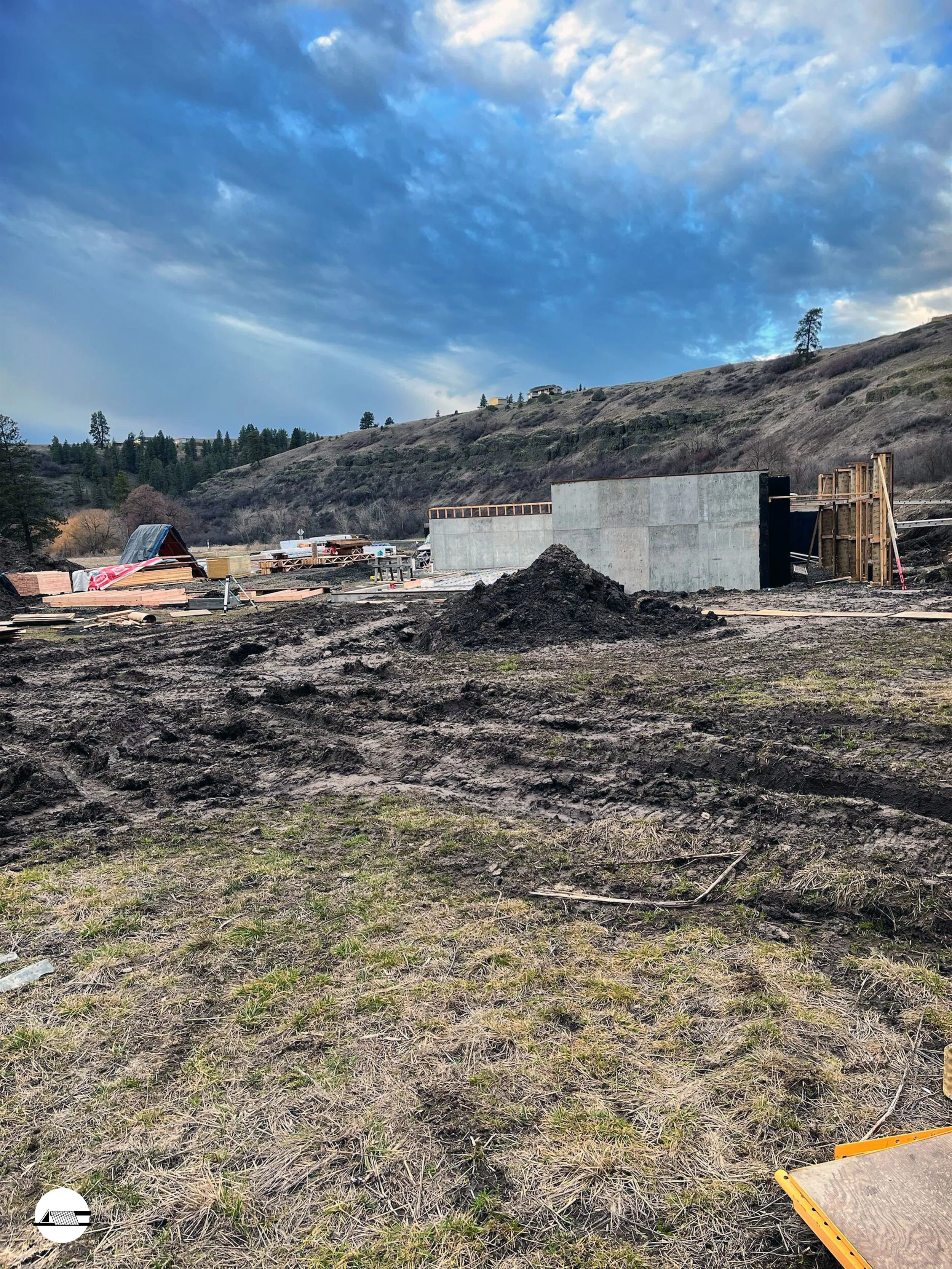 Construction site with partially built structure, dirt ground, and cloudy sky in a hilly area with sparse trees.