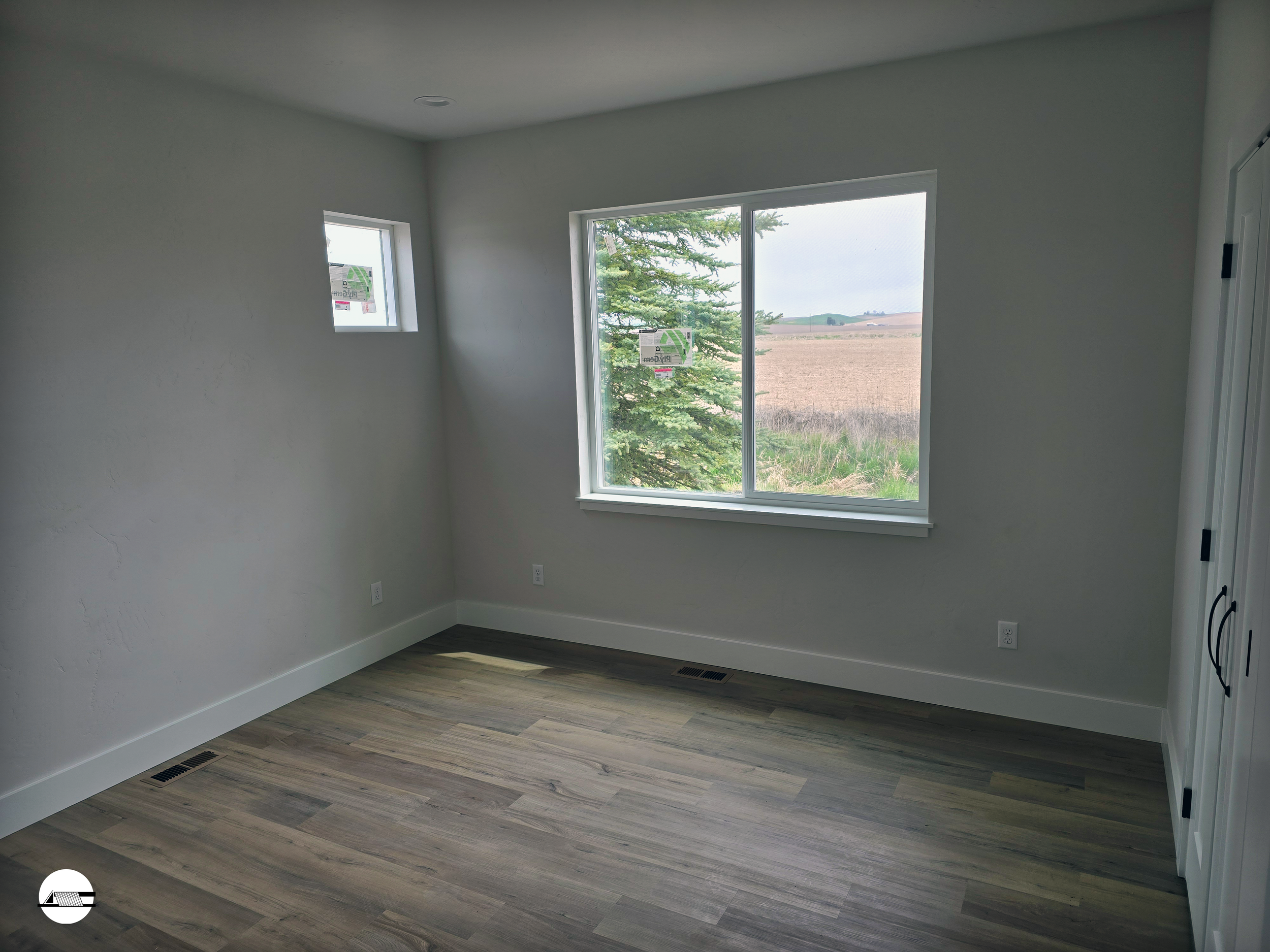 An empty room with white walls, hardwood flooring, a large window showing an outdoor field, and two small windows near the ceiling.