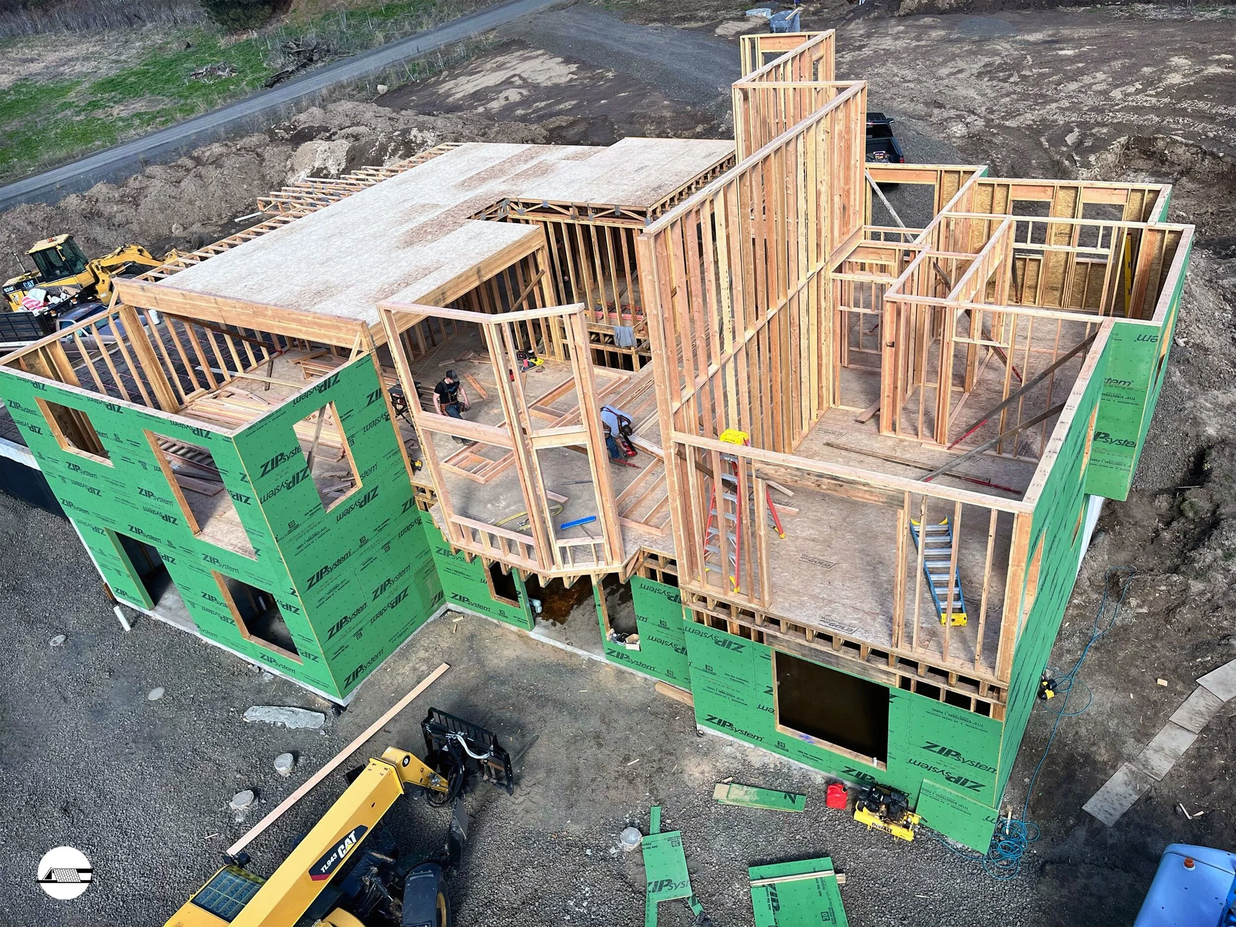 An aerial view of a house under construction showing wooden framing and green exterior sheathing. The foundation is visible with construction tools and machinery around.