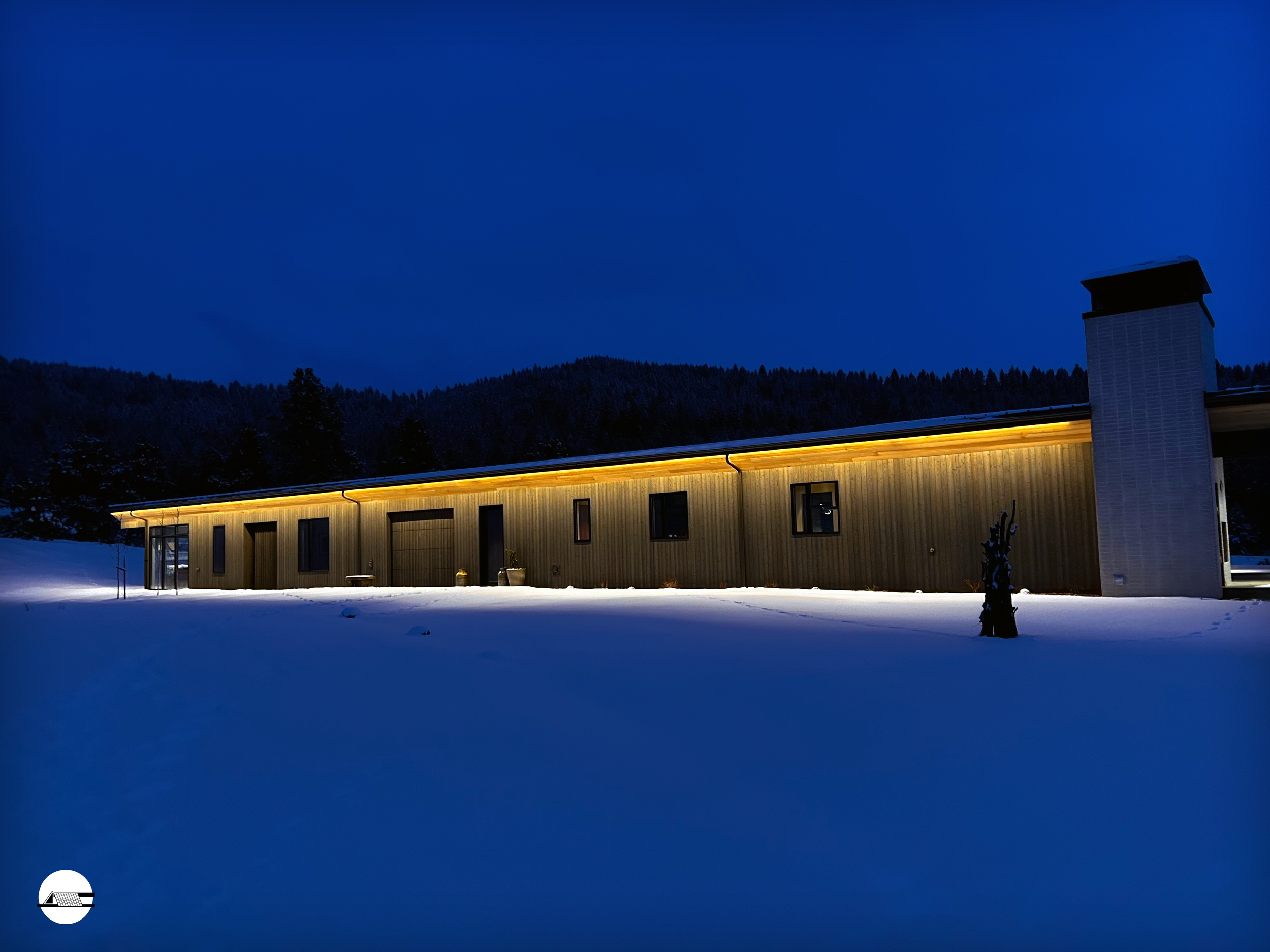 A long, single-story house illuminated with warm lights, surrounded by snow at night, with a dark forest and mountains in the background.
