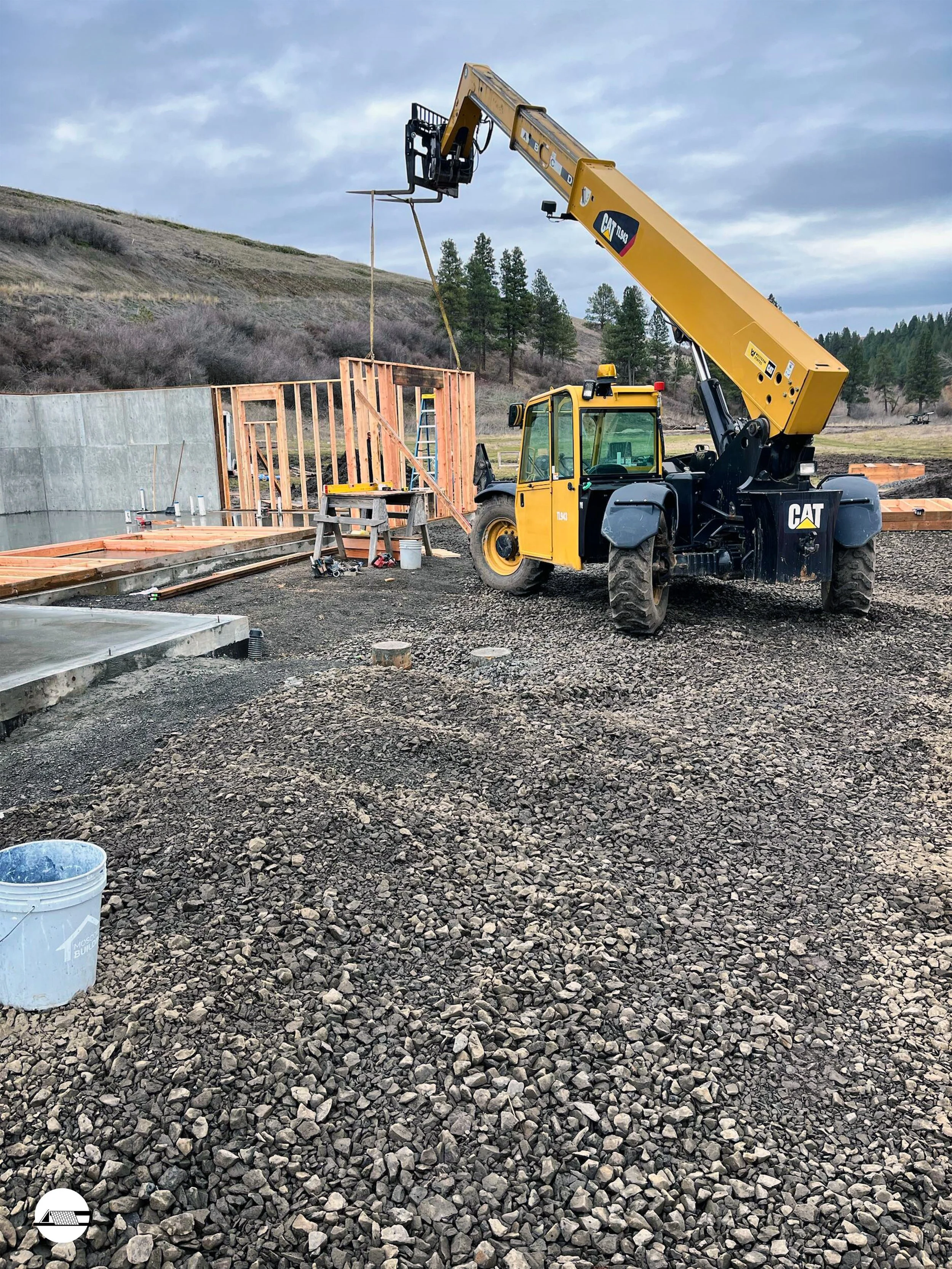 Construction site with a yellow crane lifting a wooden frame wall, surrounded by gravel ground, and partially built structures in the background under cloudy sky.