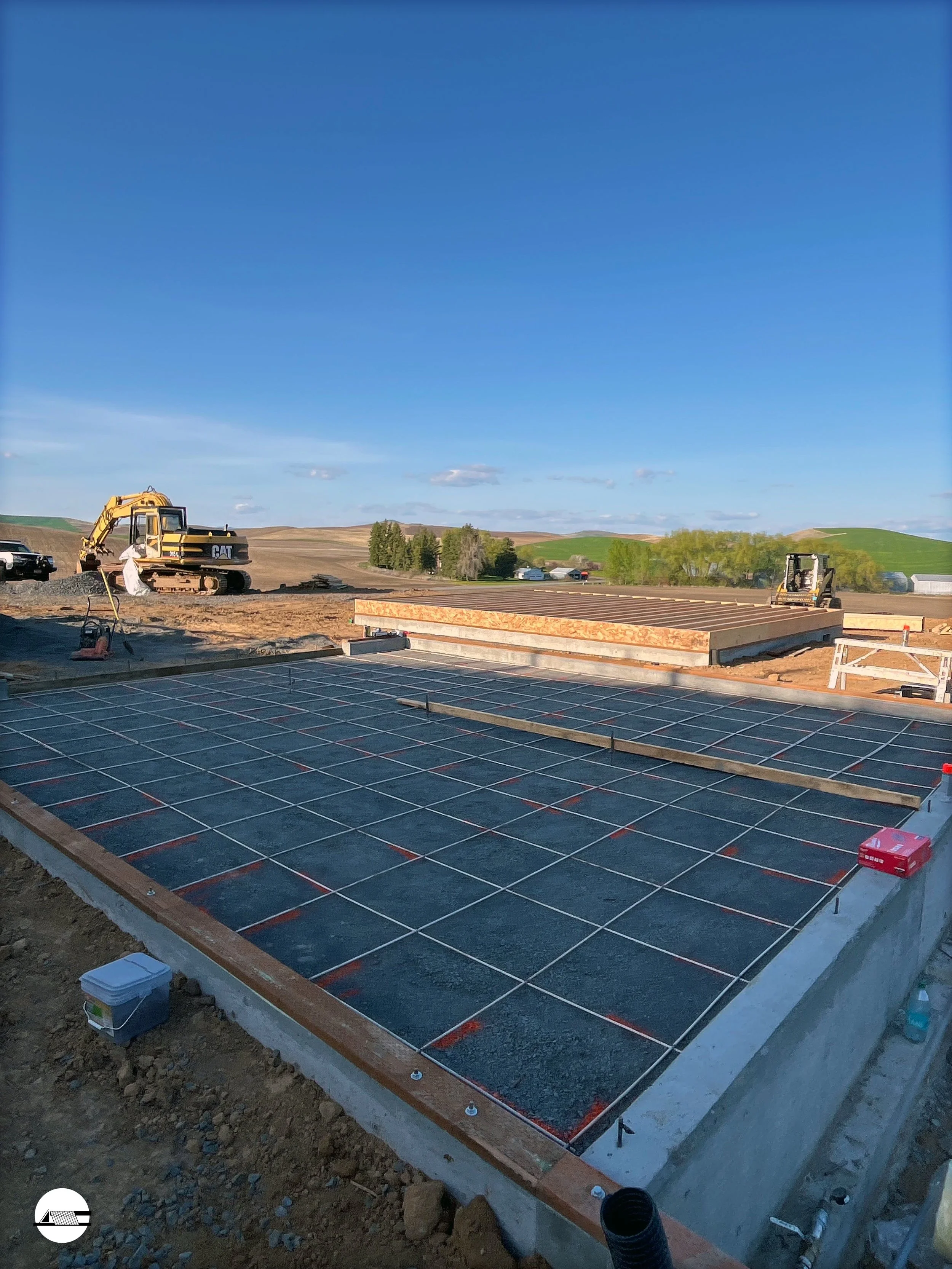 Construction site with foundation and concrete slab, wooden beams, and construction equipment like an excavator under a clear blue sky.