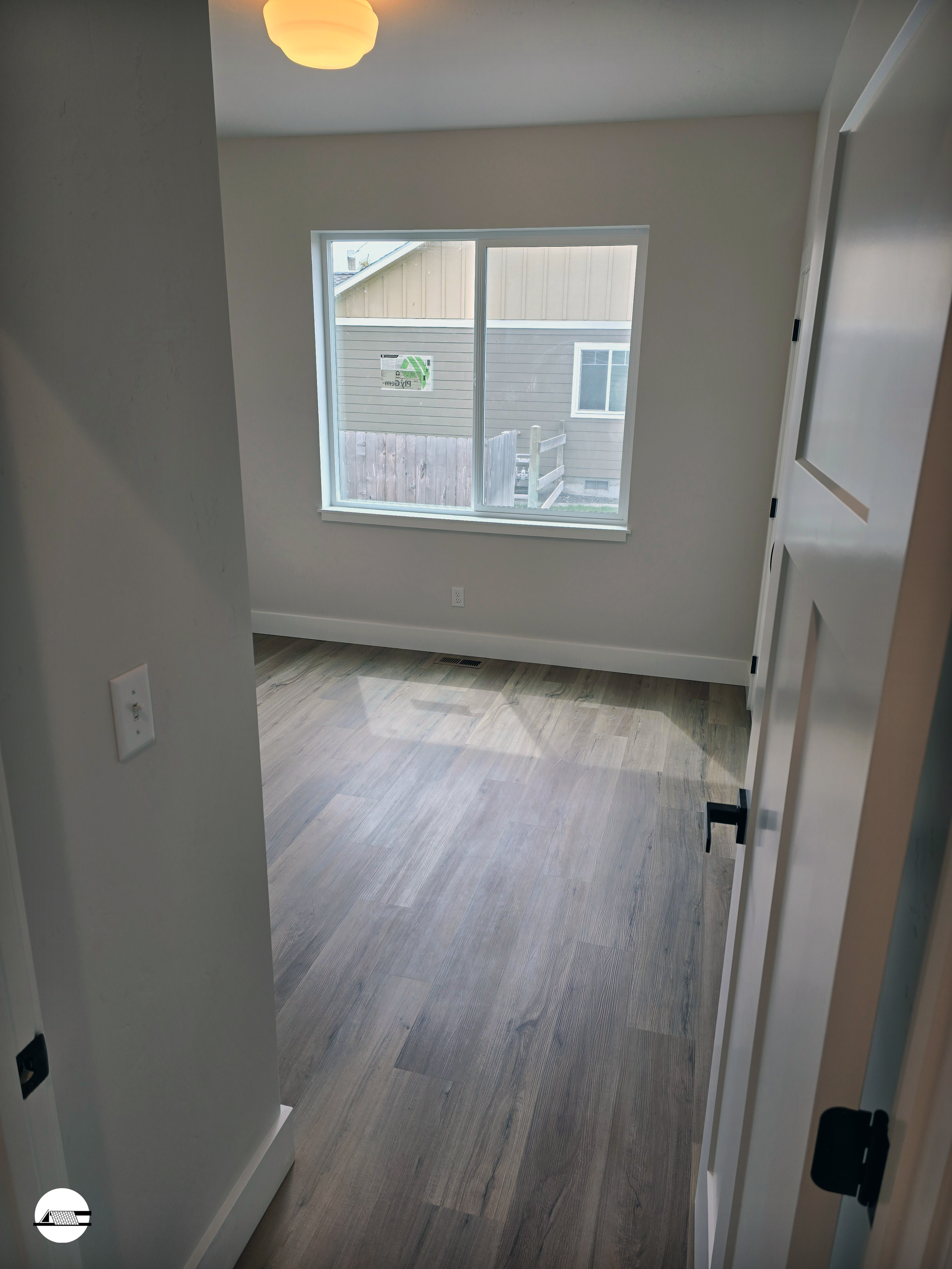 Empty room with wooden flooring, white walls, a window showing neighboring houses, and a ceiling light fixture.