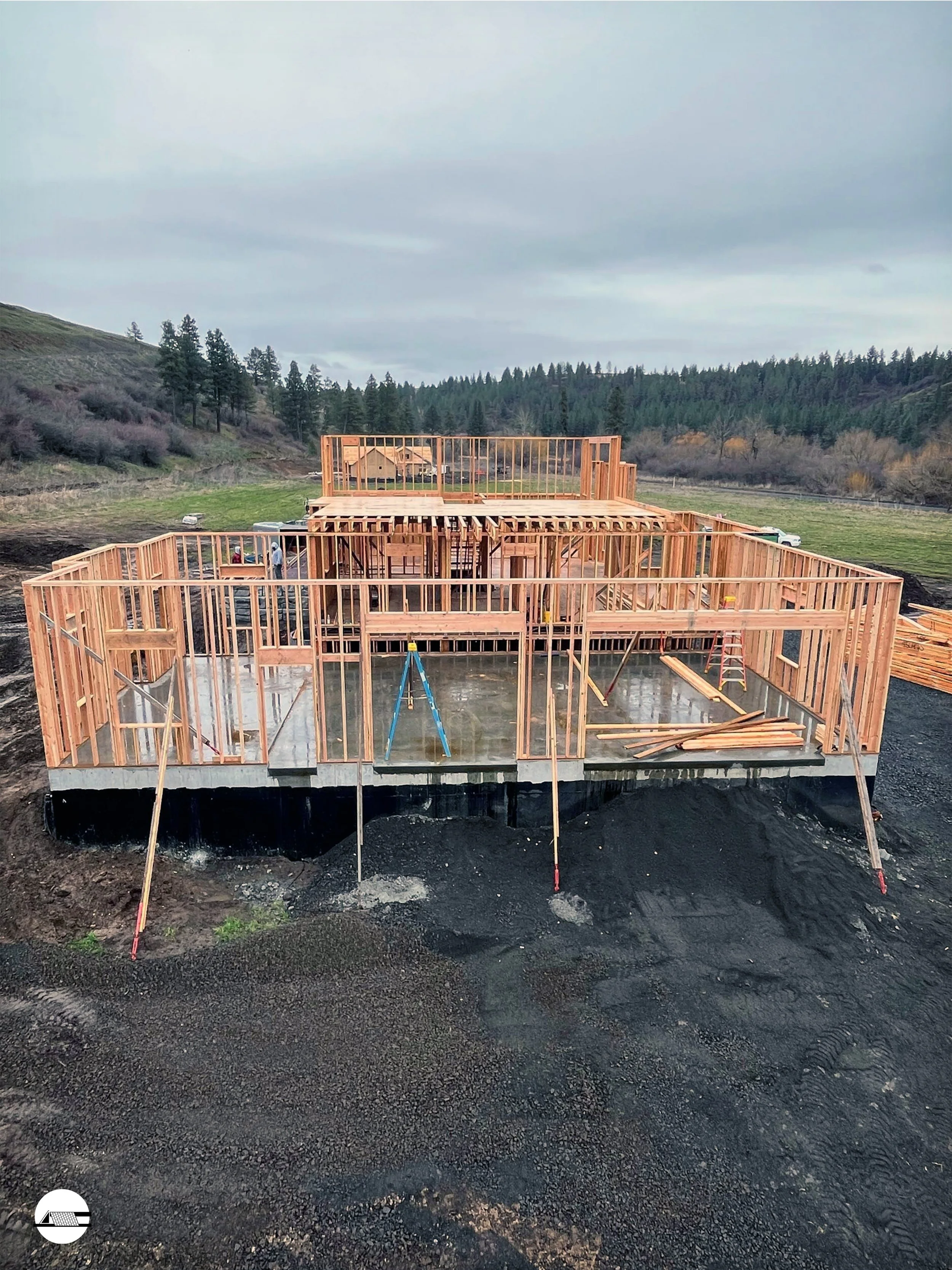 The wooden frame of a house under construction on a raised concrete foundation, with a hilly landscape and trees in the background.
