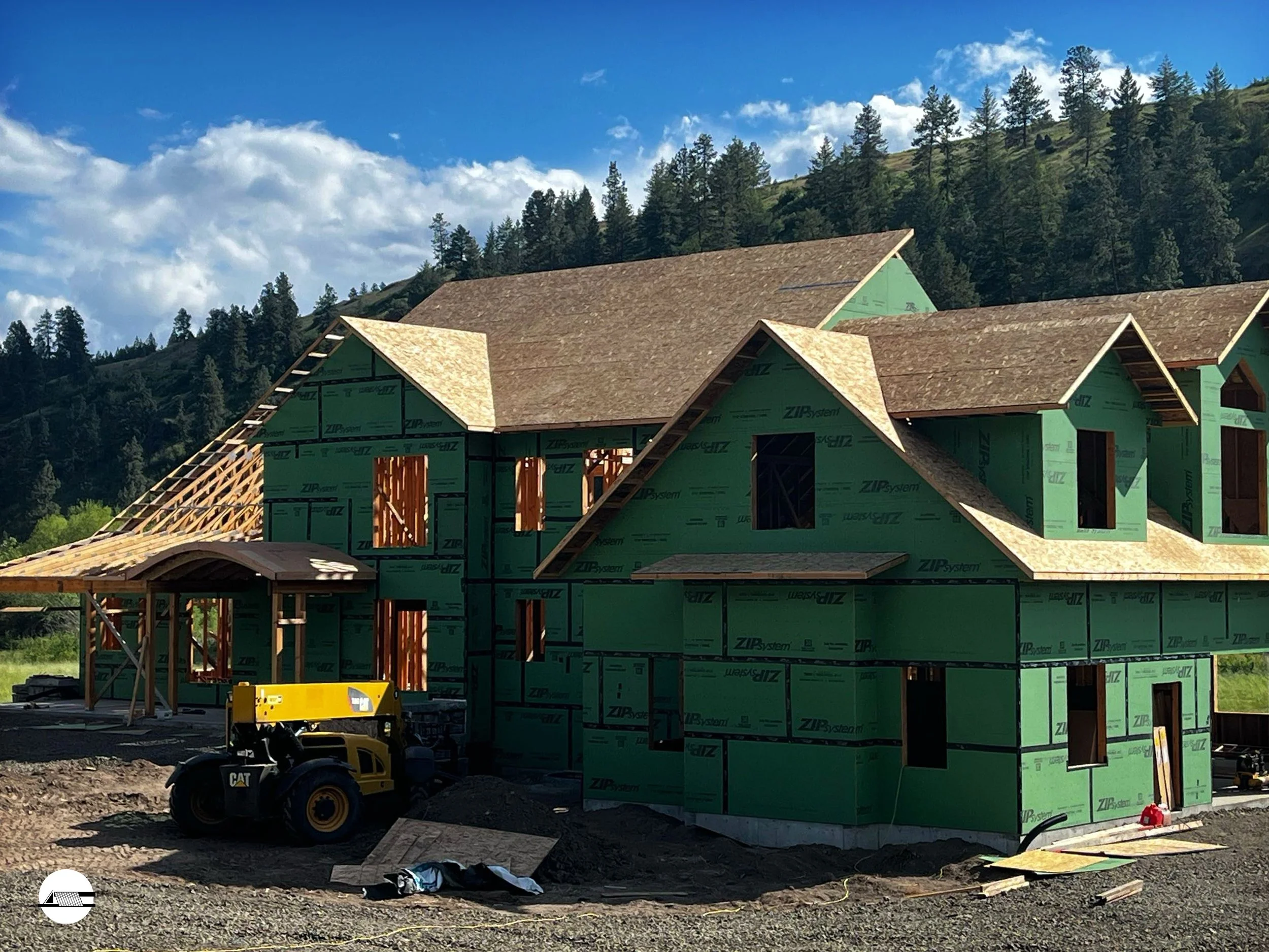A house under construction with green sheathing, wooden framing, and a sloped roof in a scenic, forested hillside setting.