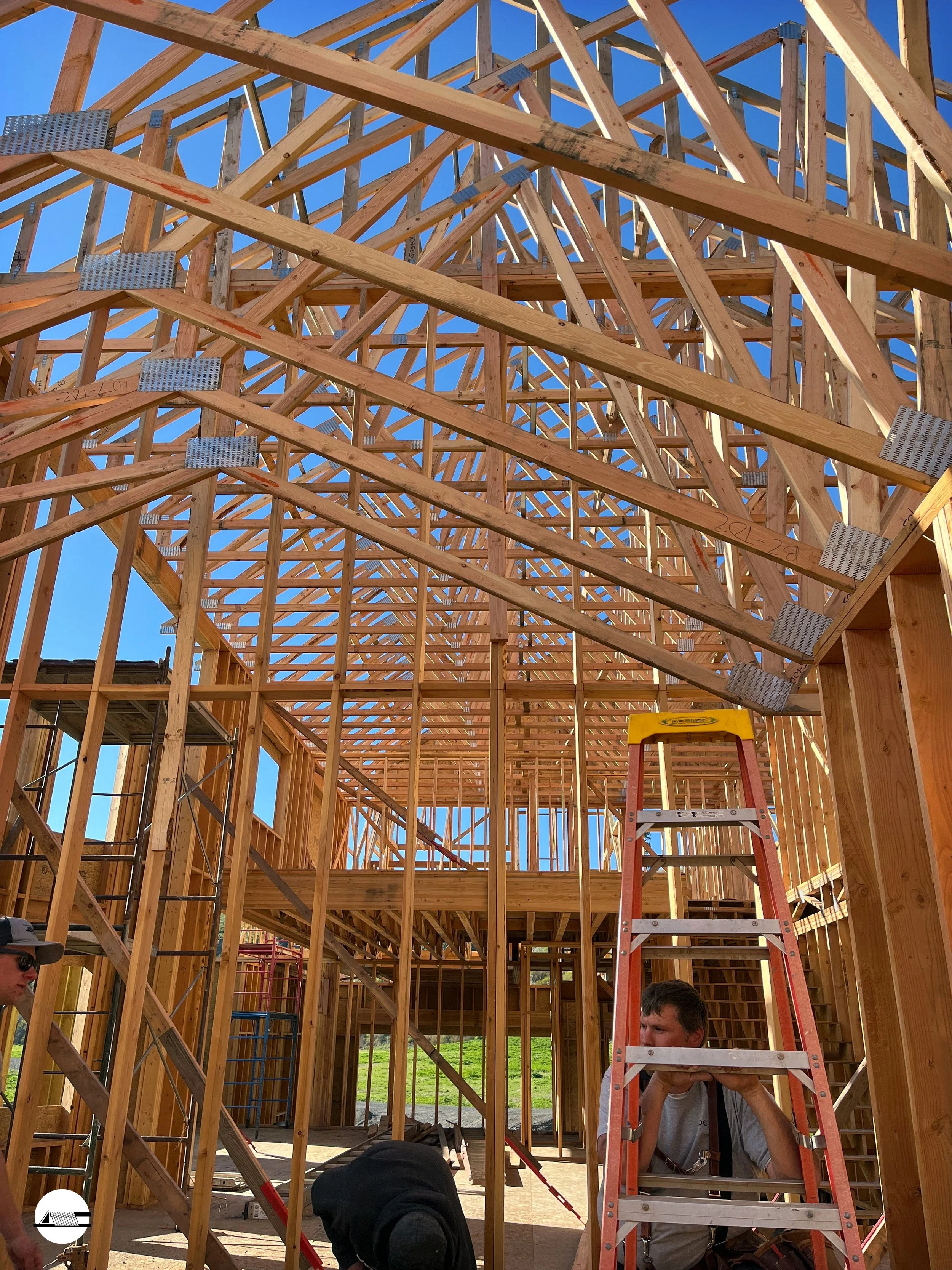 Wooden framing of a house under construction with a ladder and workers on site, set against a clear blue sky.