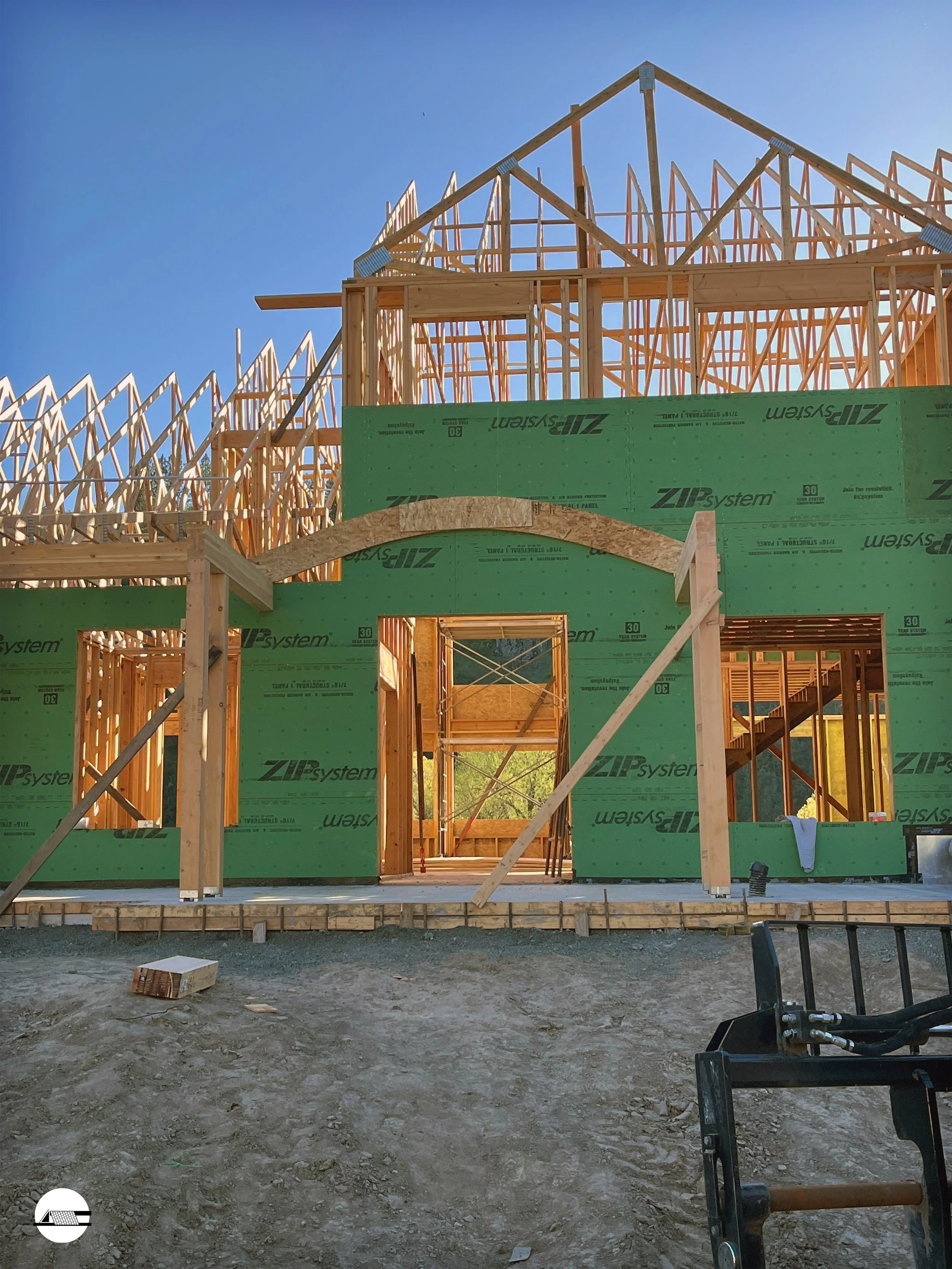Construction site of a house with wooden framing and green building wrap, under a clear blue sky.