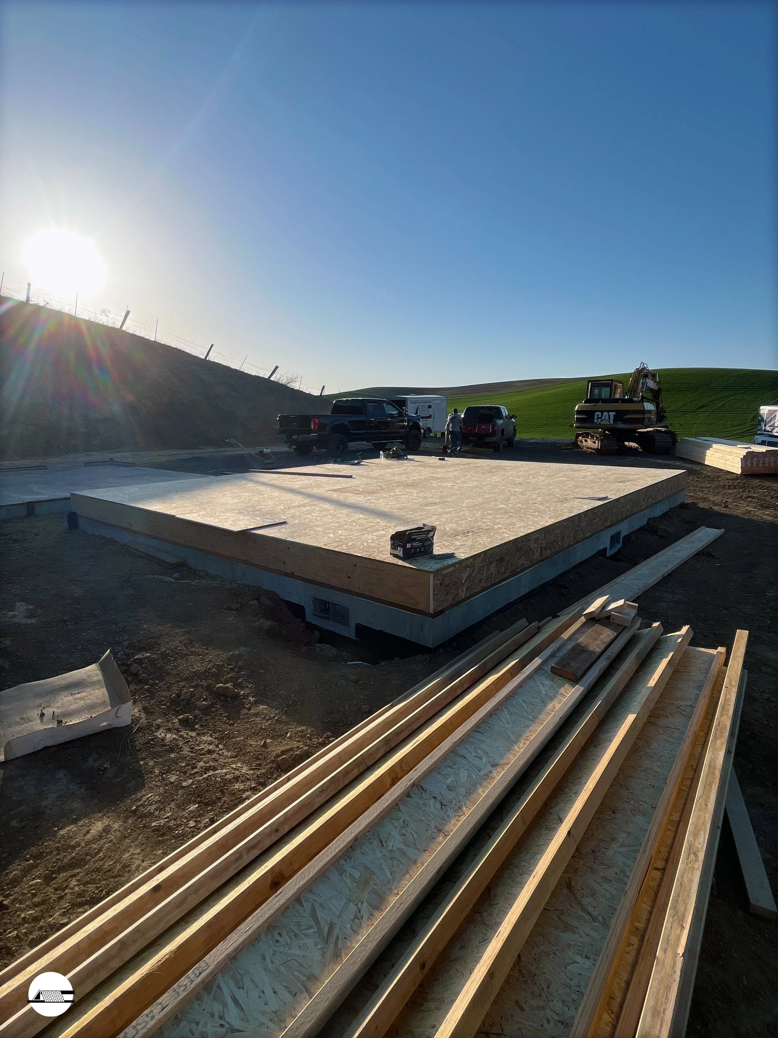 Construction site with a house foundation, wooden framing, construction vehicles, and workers working during golden hour, against a landscape of green rolling hills and a clear blue sky.