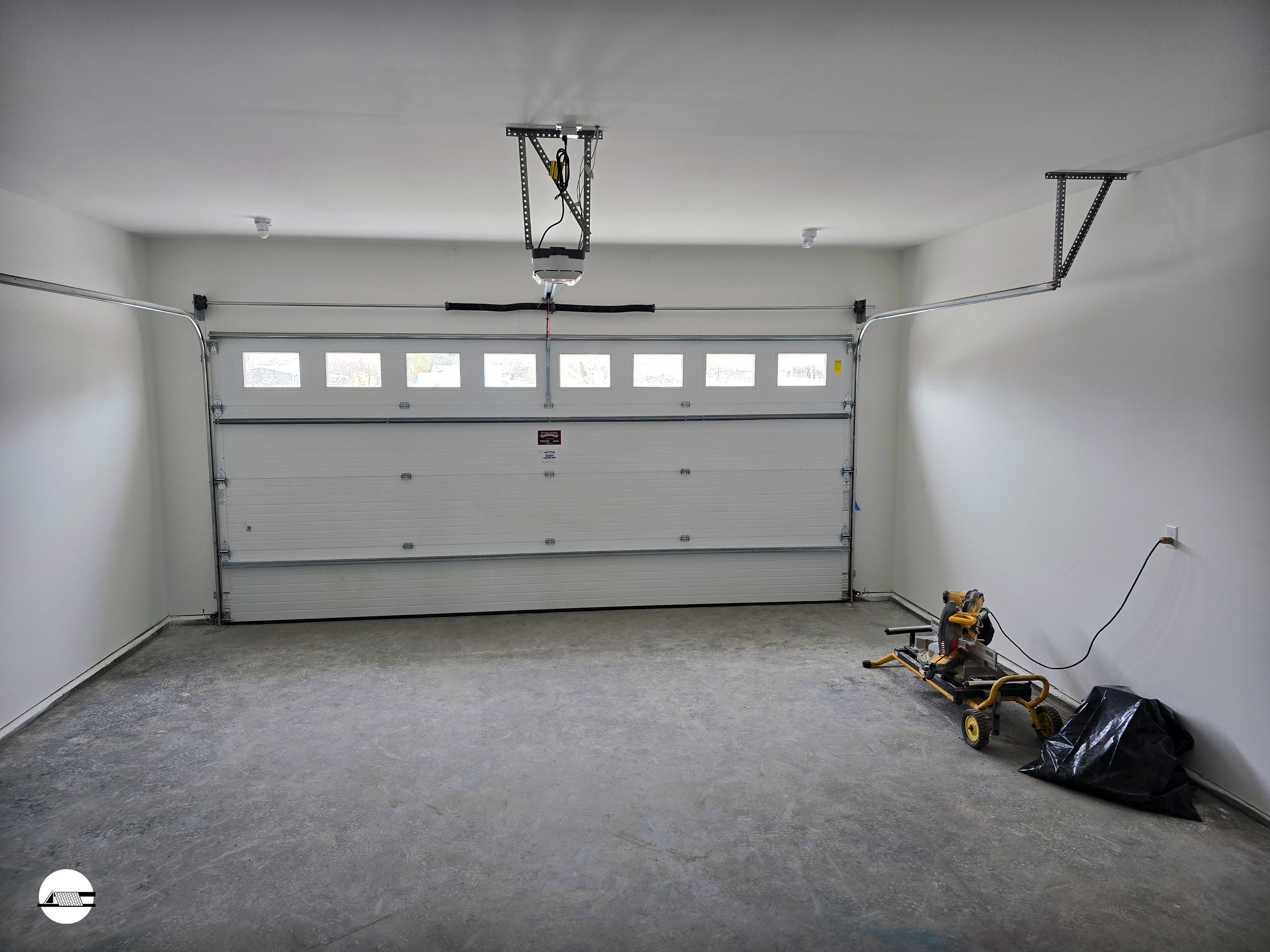 Empty garage with a closed white roll-up door and a tool on the floor, wall outlet with a cord, illuminated by natural light from small windows on the door.