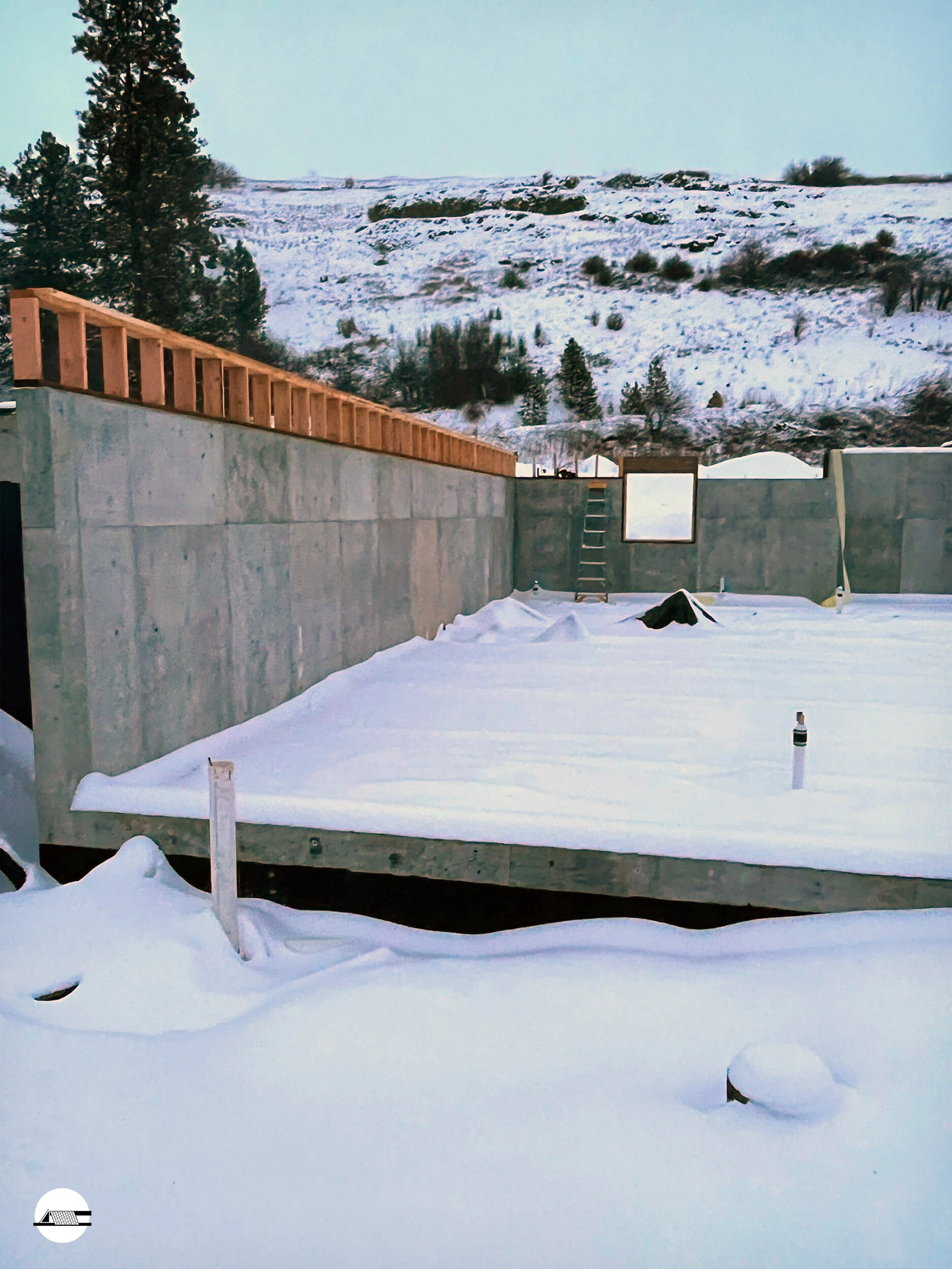 A construction site in a snowy landscape with concrete walls and wooden framing, with mountains and trees in the background.