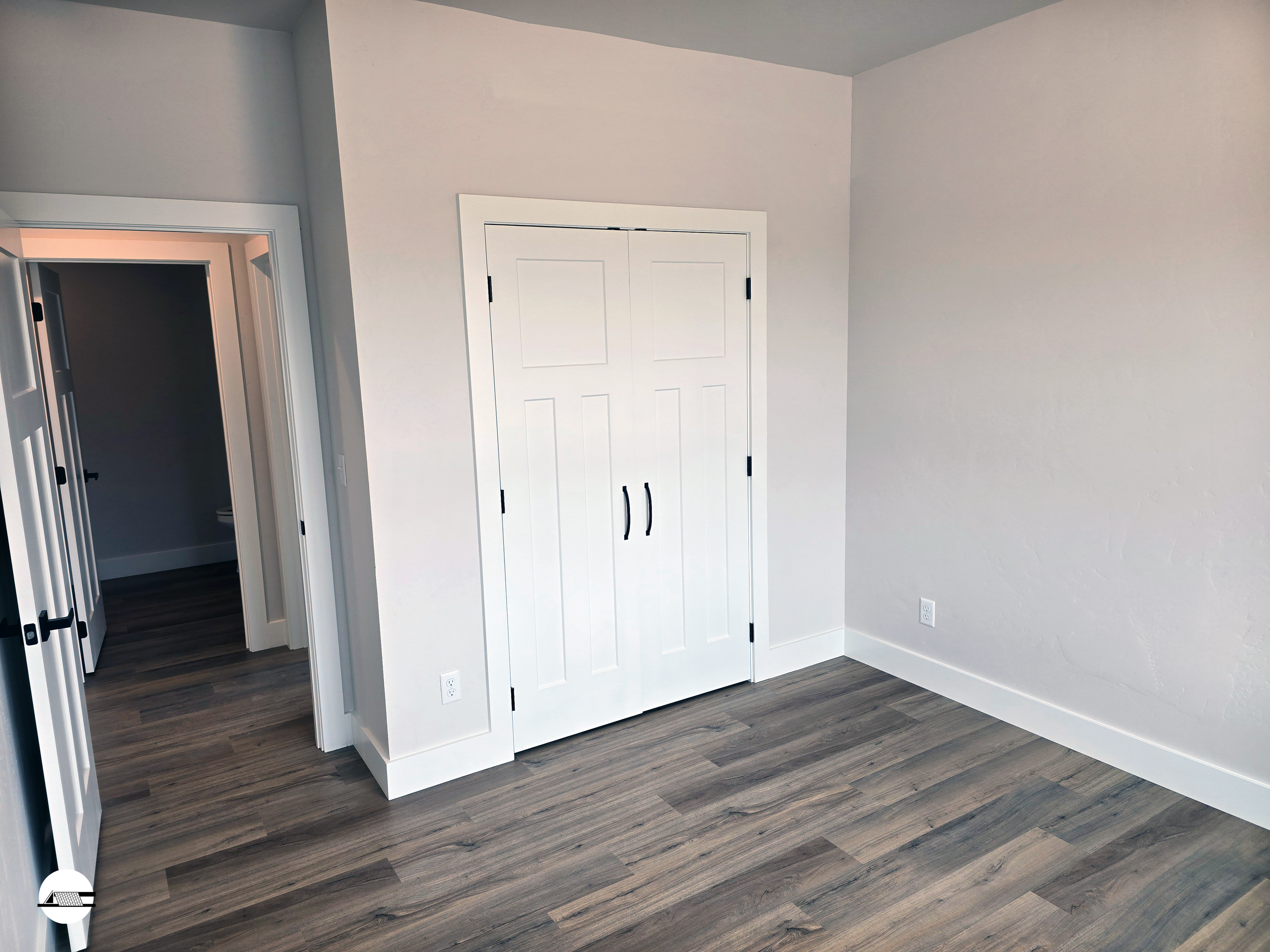 Empty room with white walls, brown wood floor, double closet doors, and an open door leading to another room.