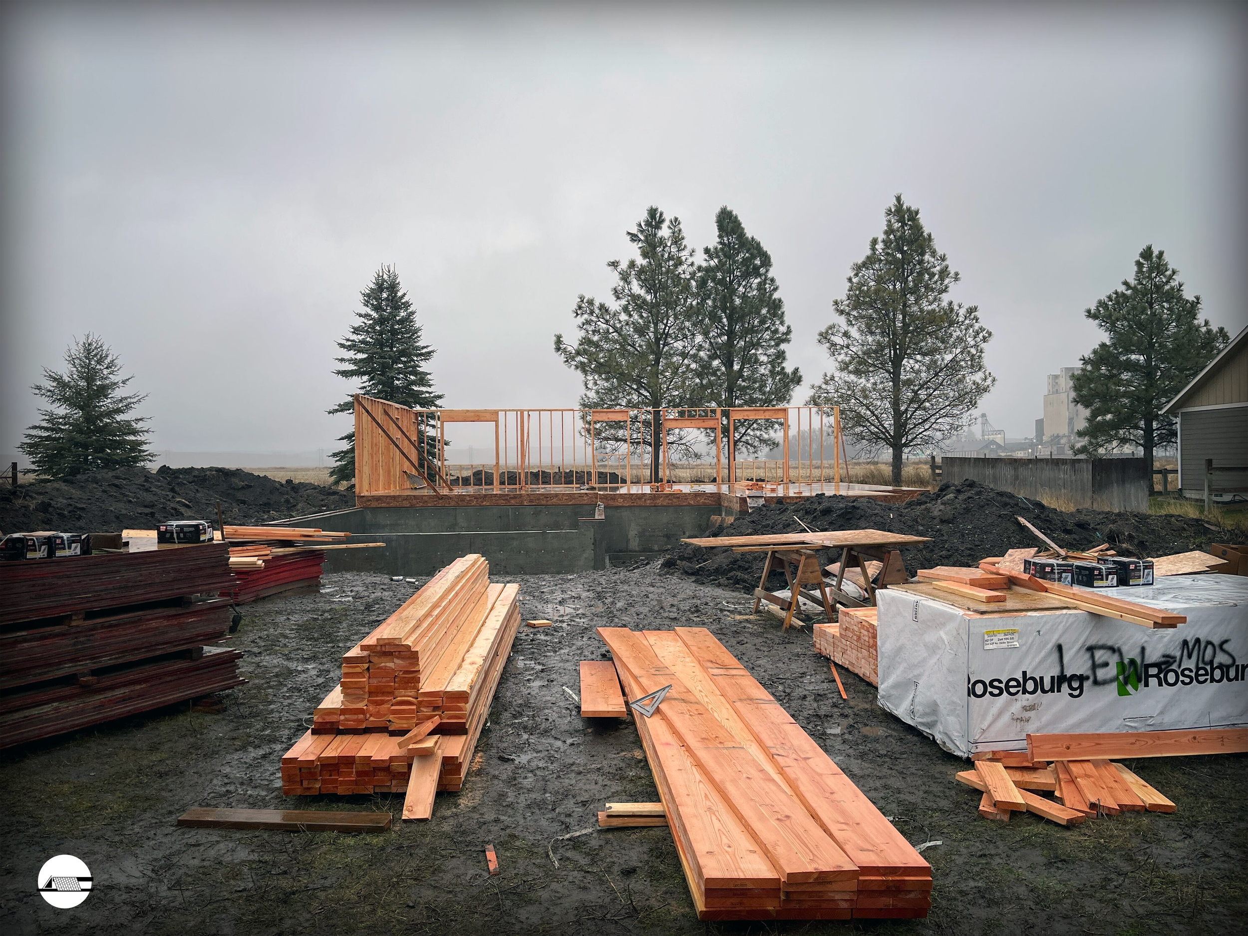 Construction site with wooden framing, lumber, and tools, set outdoors with trees and overcast sky in the background.