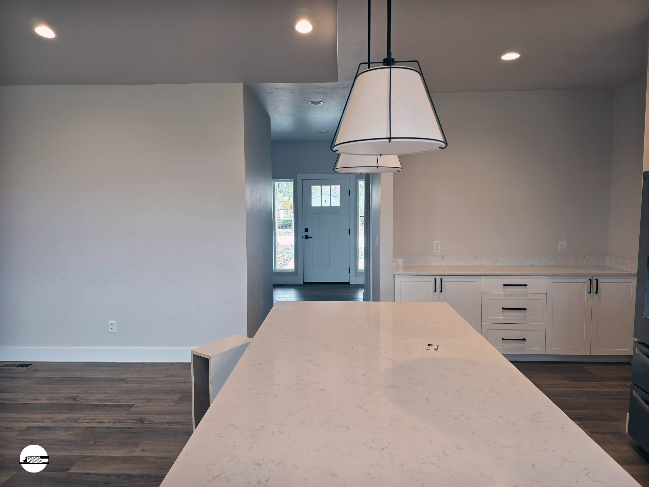 Modern kitchen with white marble island, pendant lights, white cabinets with black handles, and hardwood flooring, leading to the front door with glass panels.