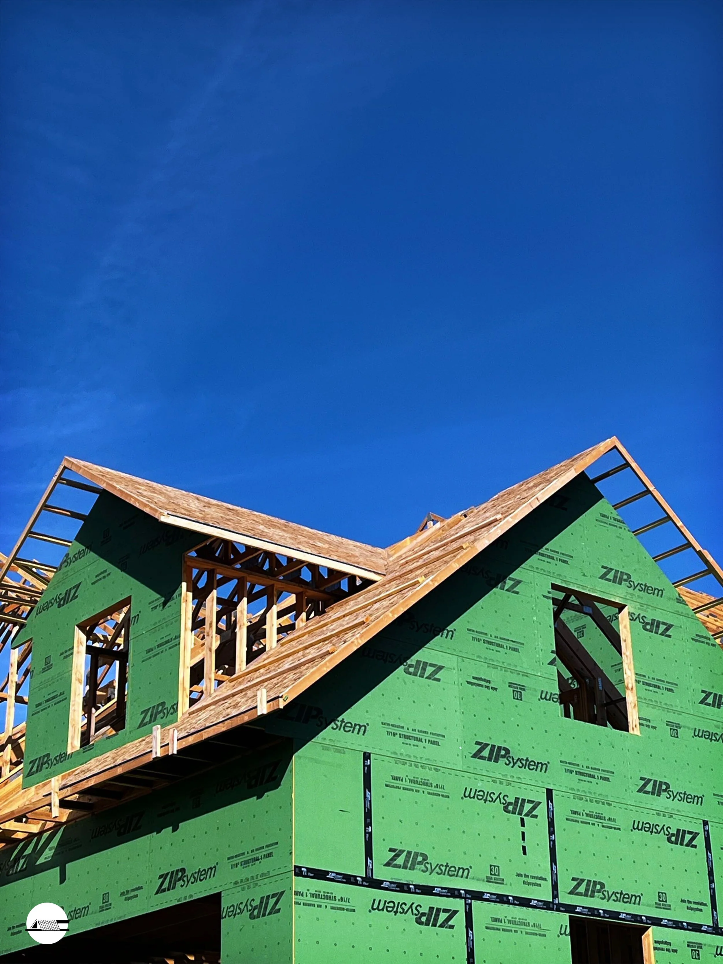 A house under construction with green ZIP system sheathing and exposed wooden framing, against a bright blue sky.