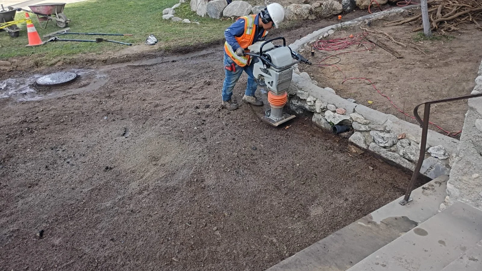 Worker compacting the ground beside a stone retaining wall.
