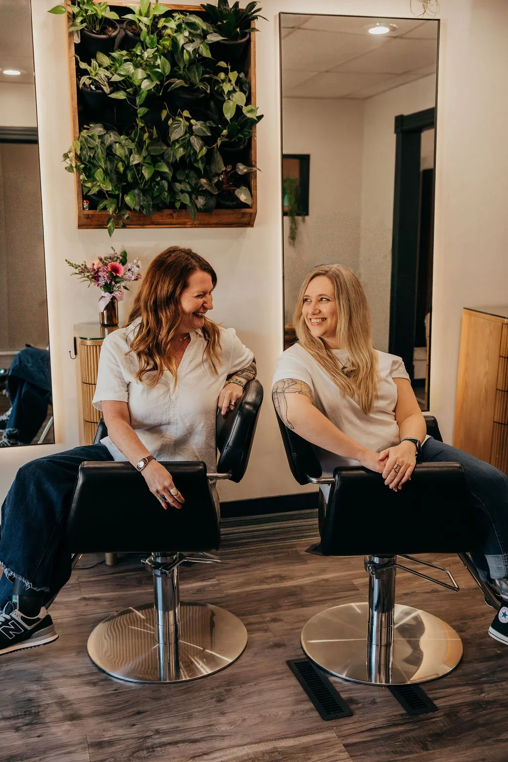 Two women are sitting in hair salon chairs, smiling and laughing at each other. One woman has long curly hair, tattoos on her arm, and is wearing a white shirt. The other woman has straight blonde hair, tattoos on her arm, and is also wearing a white shirt. Behind them, there is a wall with a mirror, a wooden shelf with a green plant, and a side table with a vase of colorful flowers.