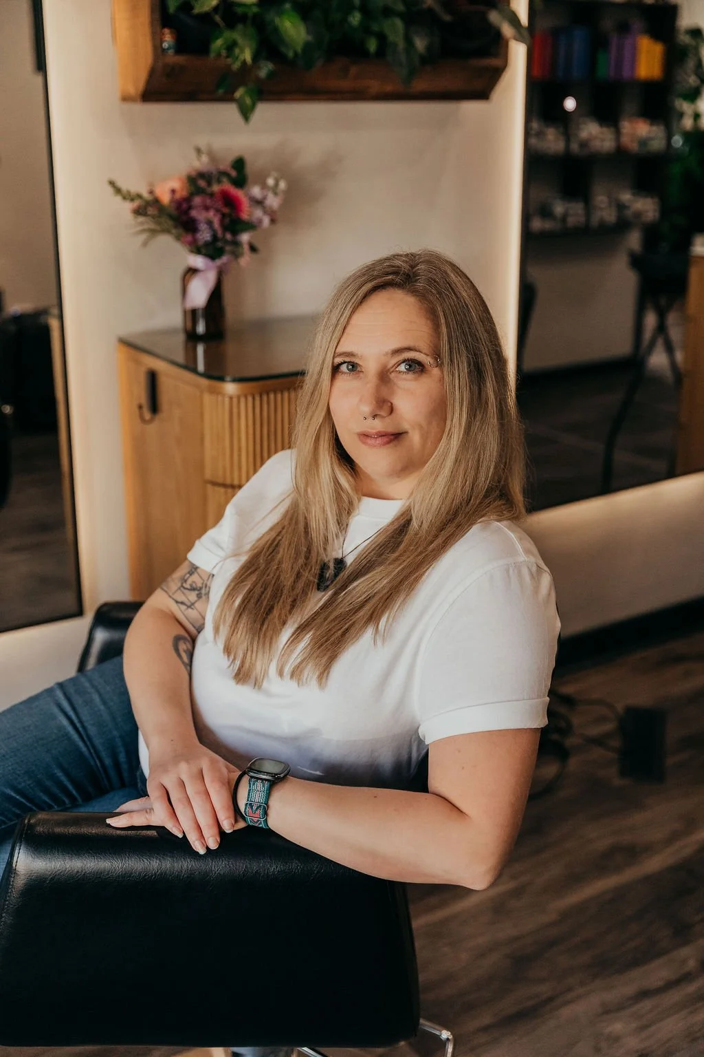 A woman with long blonde hair and a nose piercing, wearing a white t-shirt and a watch, sitting on a black chair in a cozy room with a wooden cabinet and a bouquet of flowers in the background.
